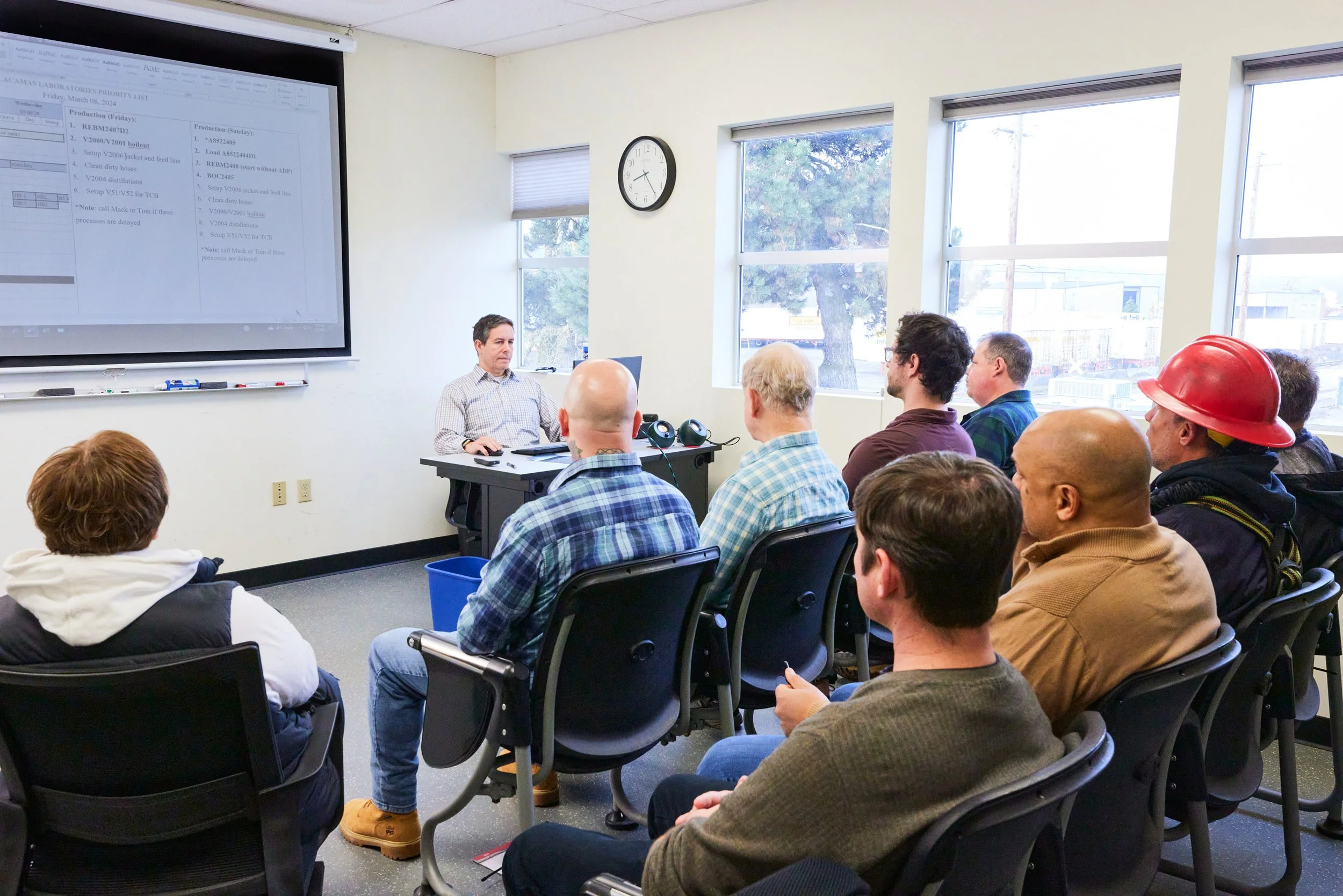 A group of people attending a training session, listening to a speaker at the front. There is a large screen displaying a presentation on the wall, and several windows letting in natural light. The presenter is seated at a desk with a laptop, and one person is wearing a red safety helmet.