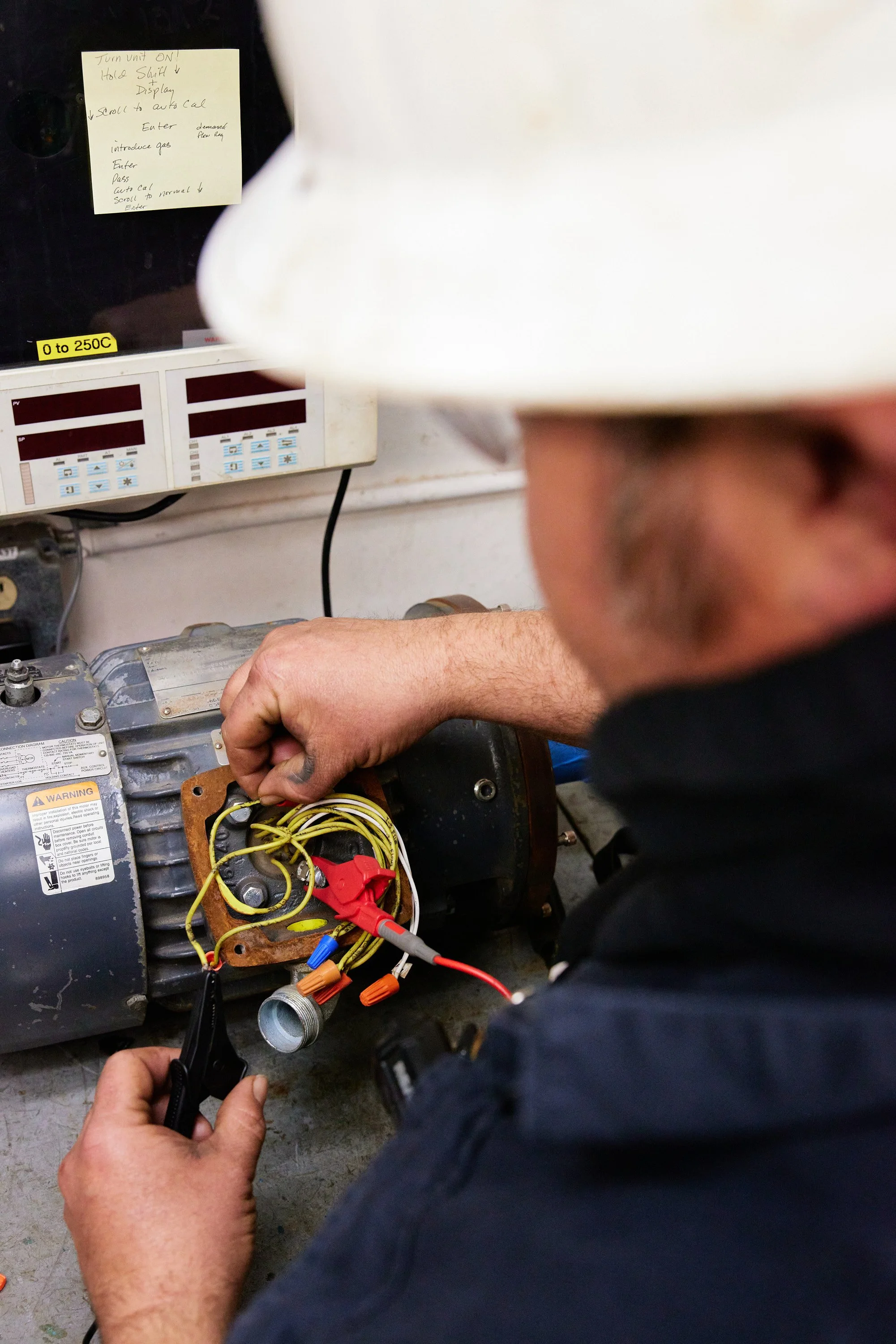 A person is working on an electrical motor, connecting wires with a multimeter. In the background, there is a control panel with instructions and temperature settings.