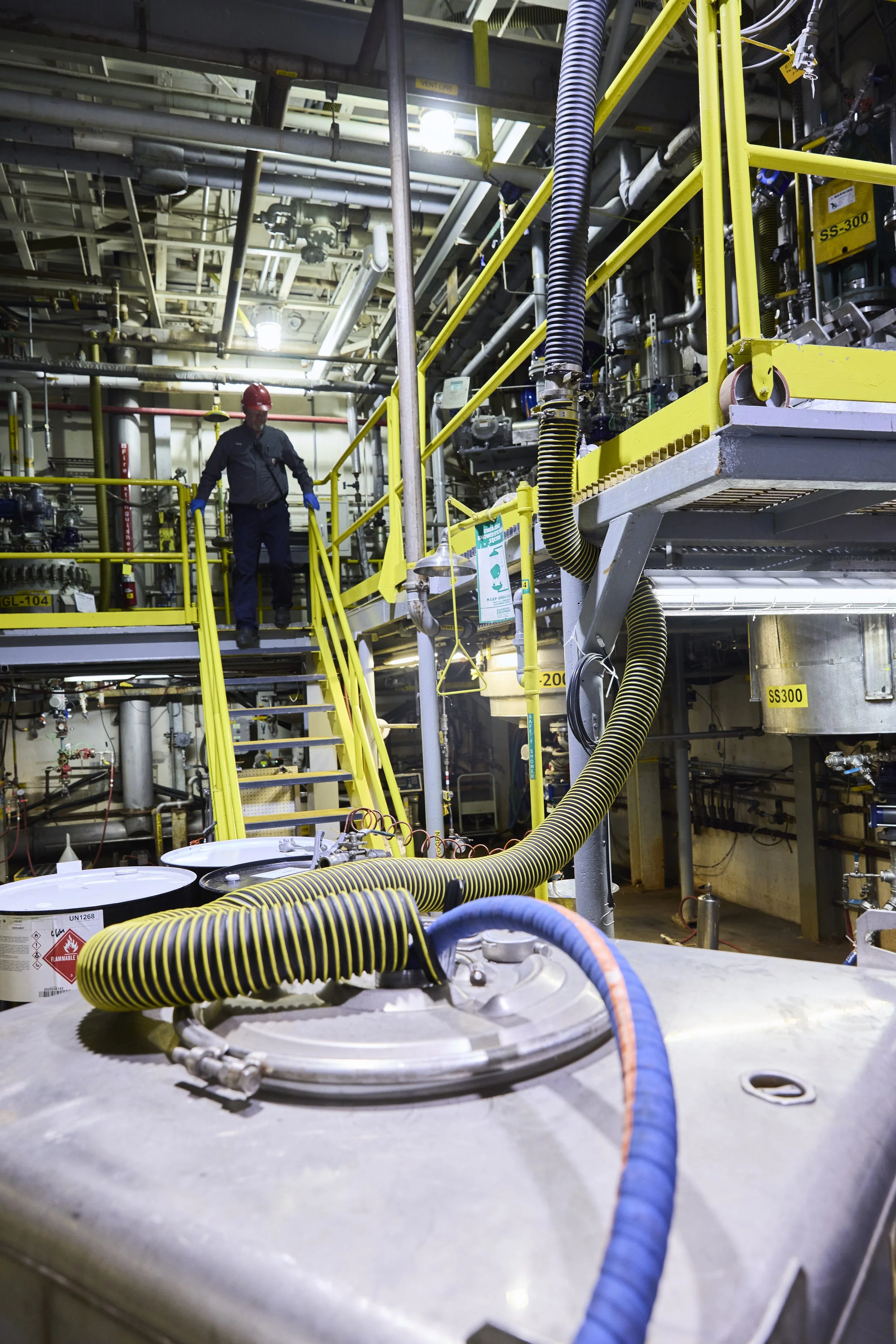 An industrial facility with yellow safety railings, pipes, and hoses. A worker wearing a red helmet and dark uniform with blue gloves is walking on a yellow staircase. The environment appears to be a manufacturing or processing plant with various equipment and safety signs.