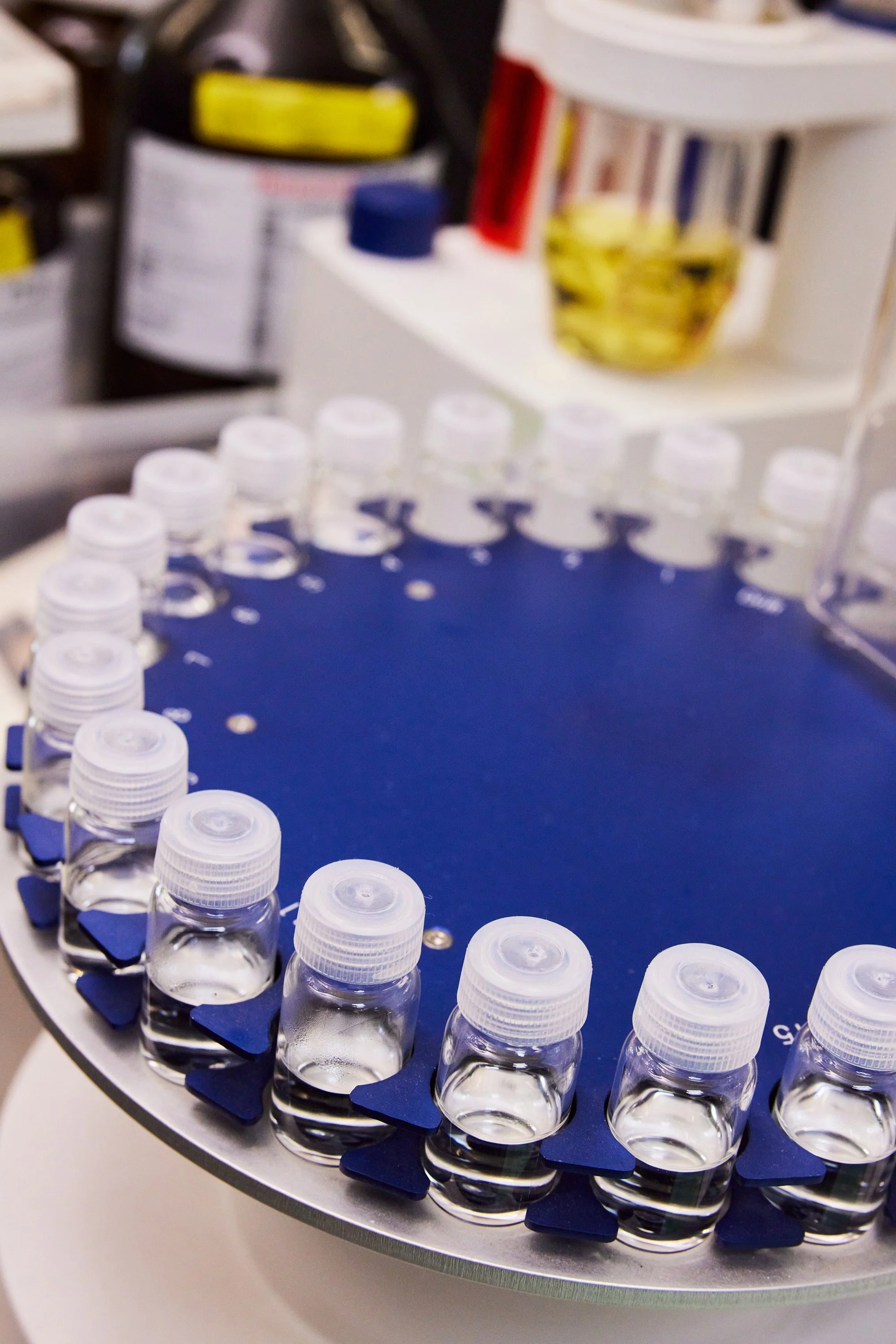 Close-up of a circular rack with small bottles of liquid, arranged in a circular pattern, with medication bottles and containers in the background.