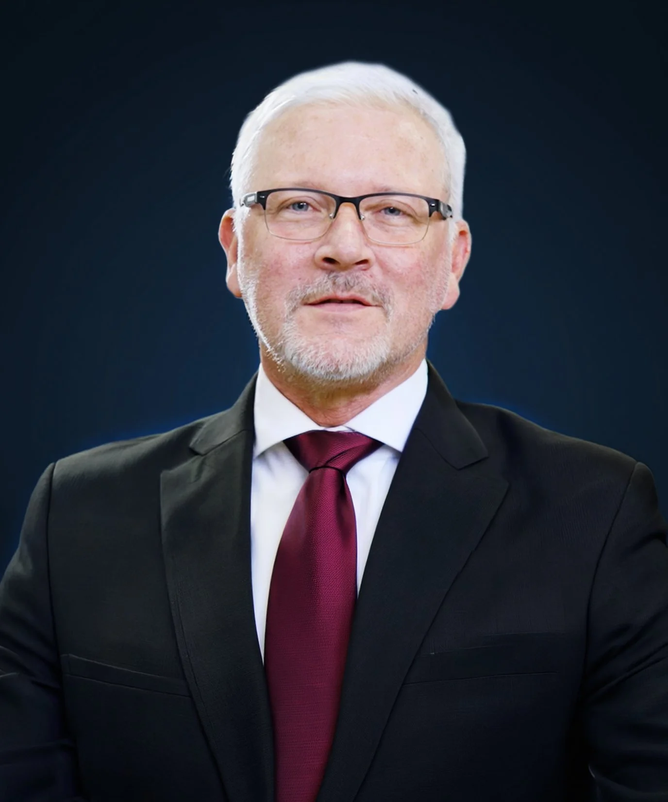 A man with gray hair, glasses, and a beard wearing a black suit, white shirt, and burgundy tie.