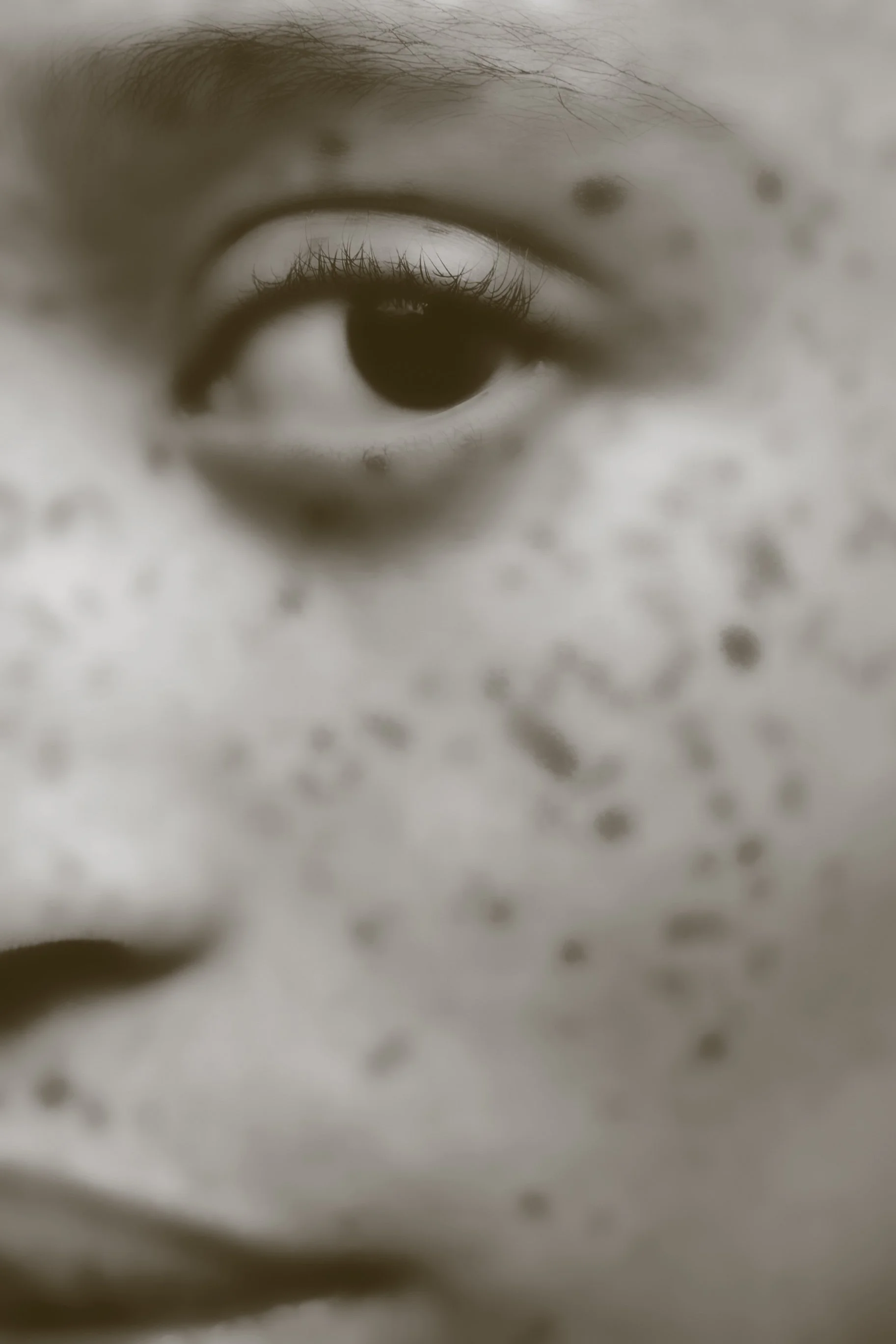 Close-up photograph of a human eye with visible eyelashes, eyebrow, and surrounding skin, in black and white.