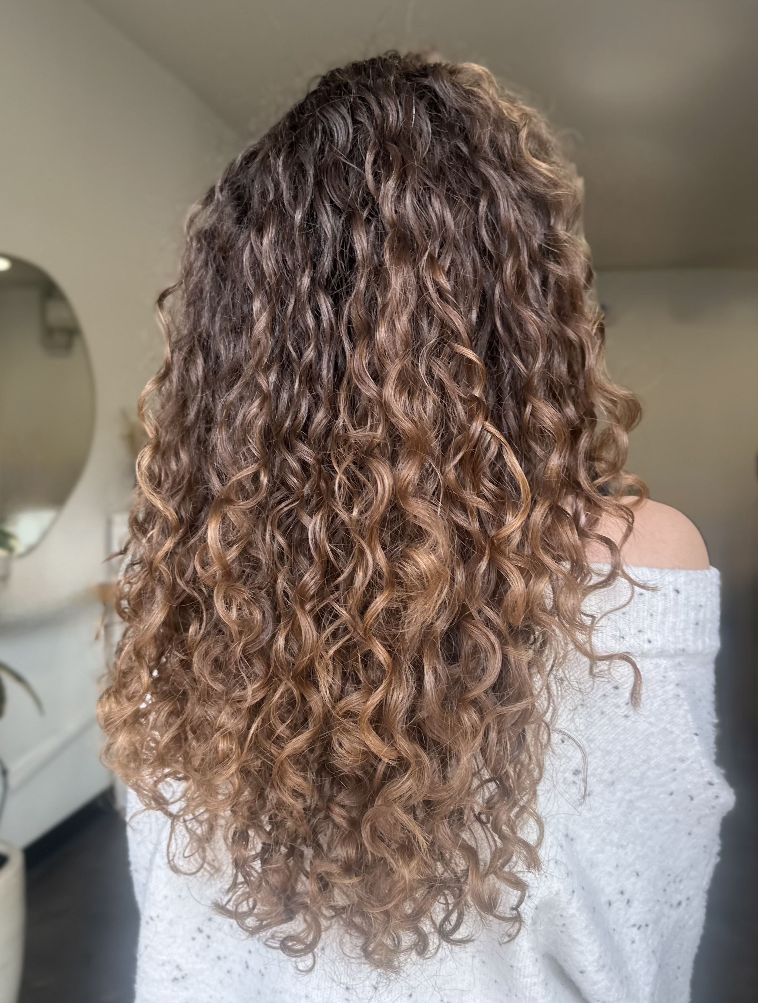 Close-up photo of long, curly, brown hair with highlights worn by a person wearing a light-colored off-shoulder top, with a blurred background including a mirror and a wall.