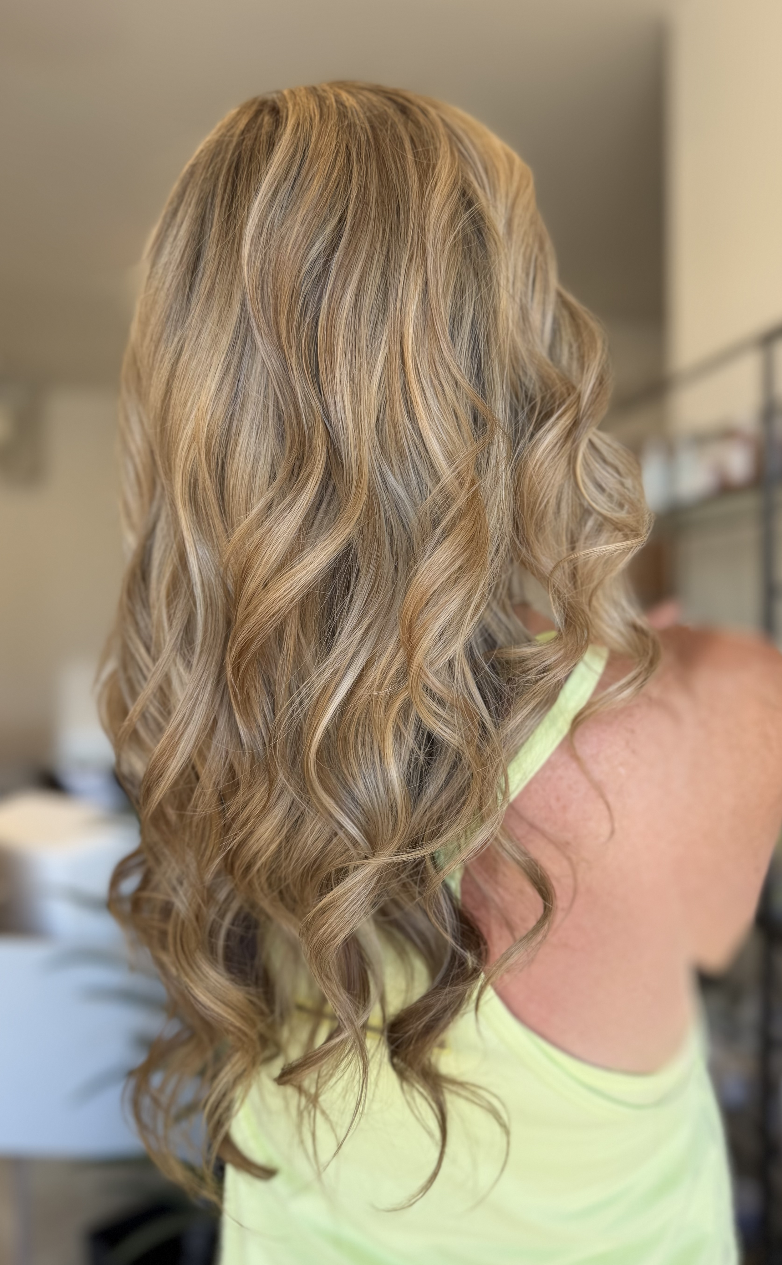 Back view of a woman with long, blonde, curly hair wearing a yellow top.