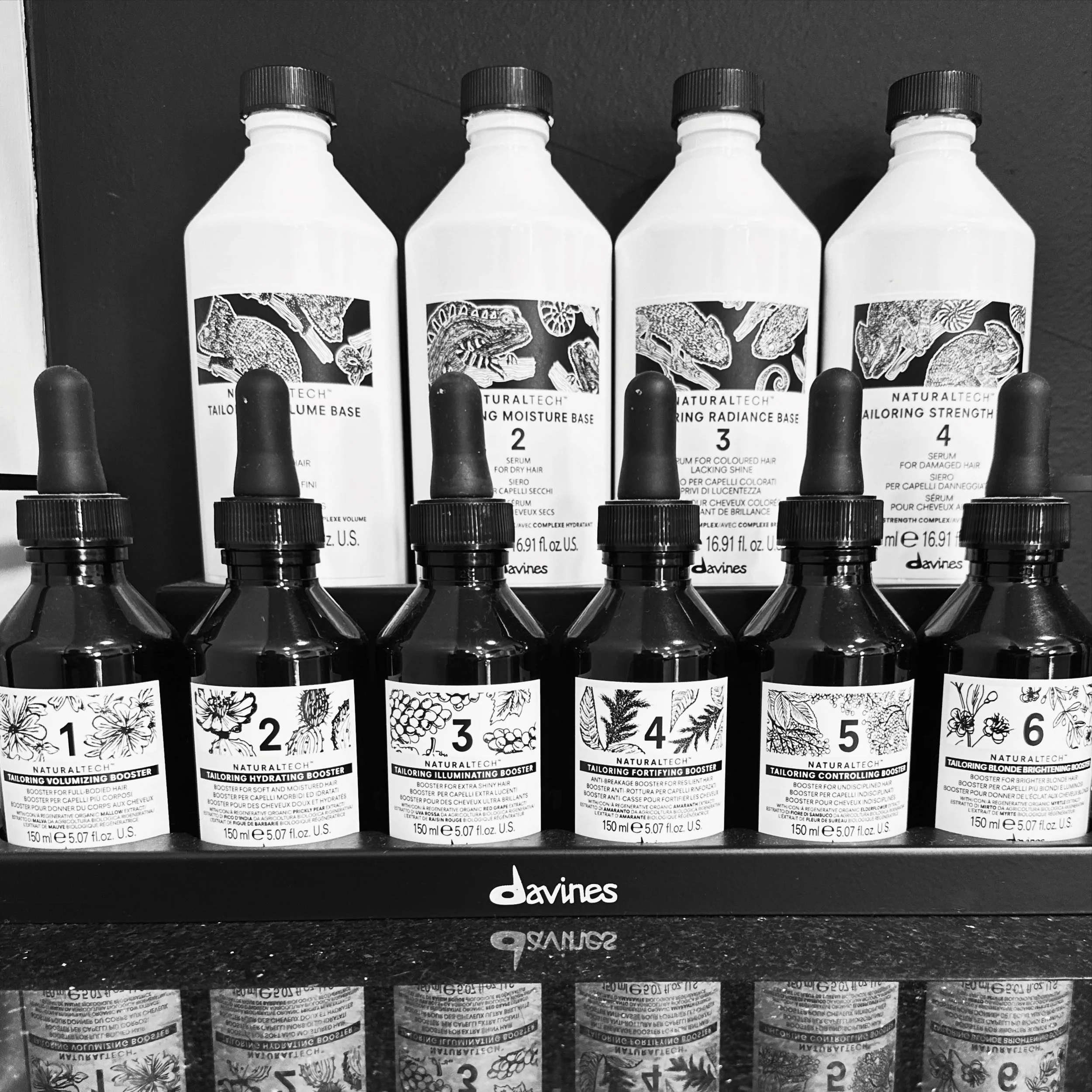 Black and white photo of four bottles of hair care products with labels featuring botanical illustrations, placed on a shelf with black pegs and a Davines logo below.