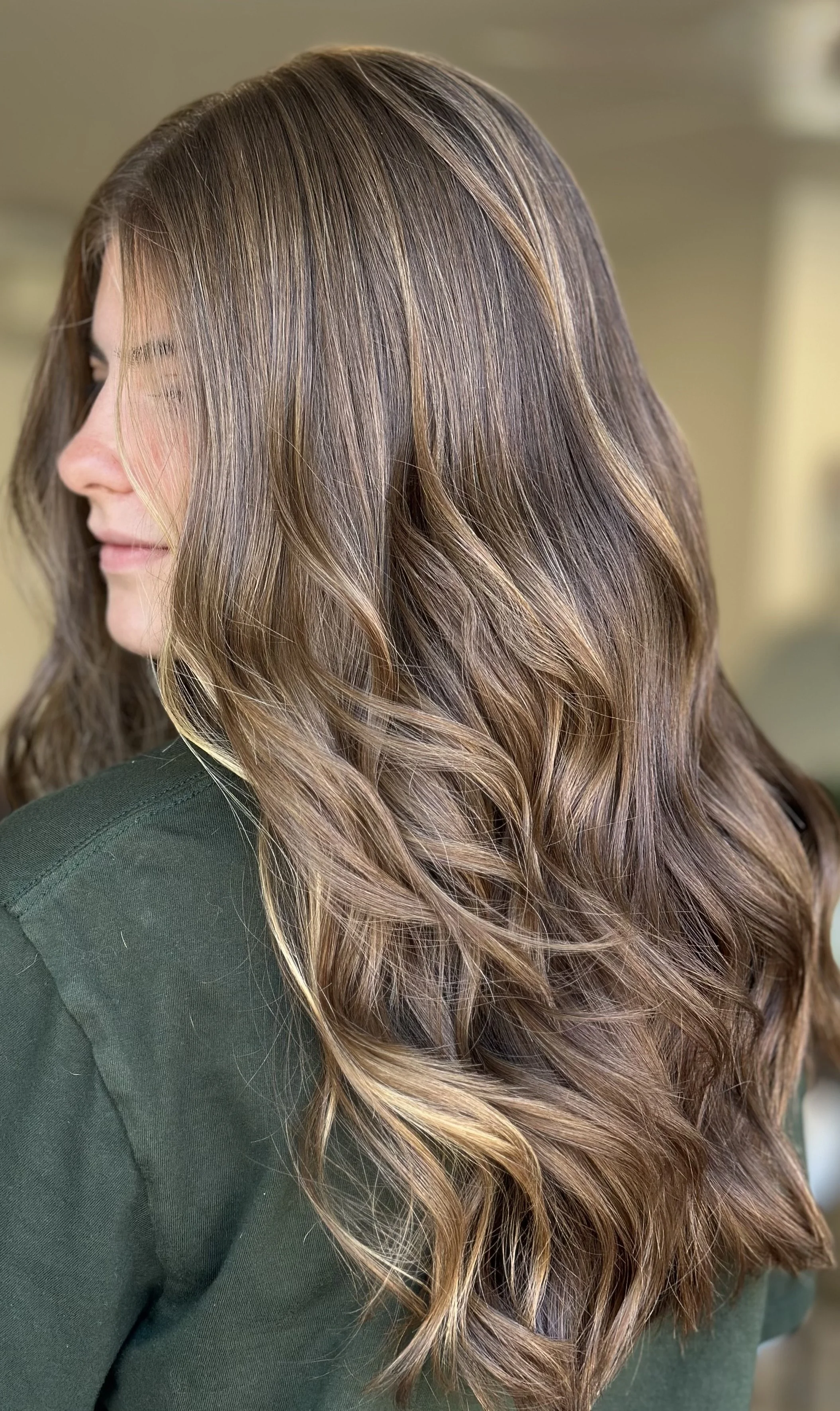 Close-up of a woman's long, wavy brown hair with subtle highlights, wearing a dark green top.