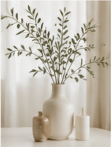 A cream-colored vase with green leafy branches, a small beige vase, and two white candles on a white surface.