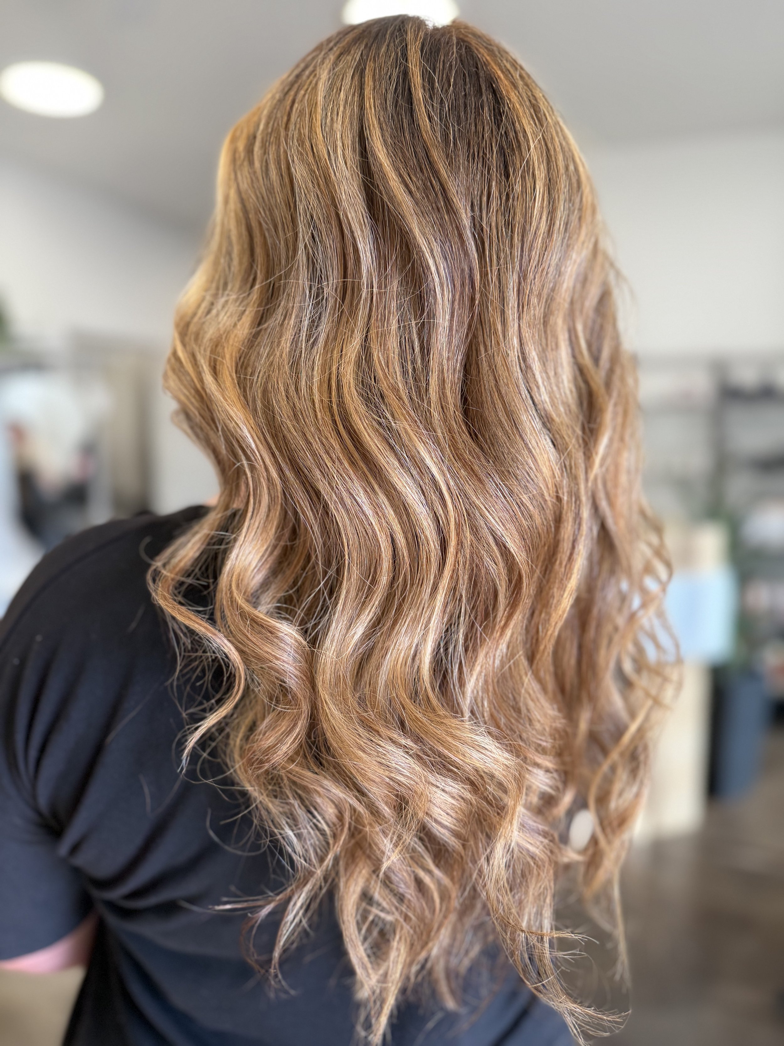 Back view of a woman with long, wavy, multi-tone hair in blonde and light brown hues, wearing a black top in an indoor setting.
