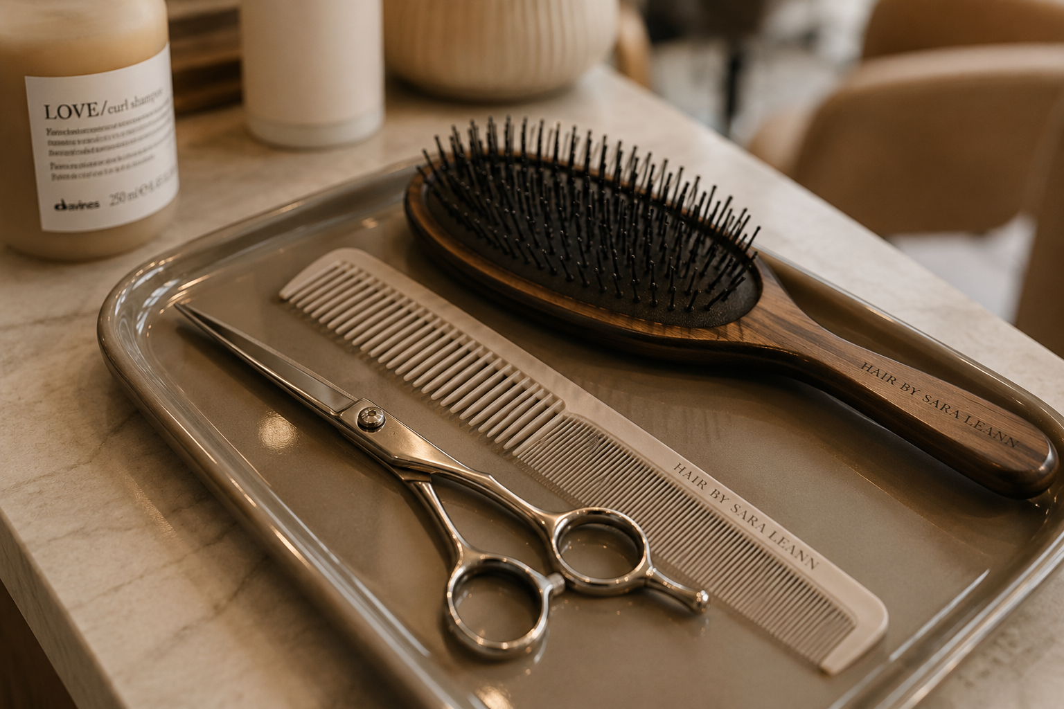 A tray holding hairstyling tools including scissors, a wide-tooth comb, a fine-tooth comb, and a wooden paddle brush with black bristles. In the background, there are bottles of hair products.