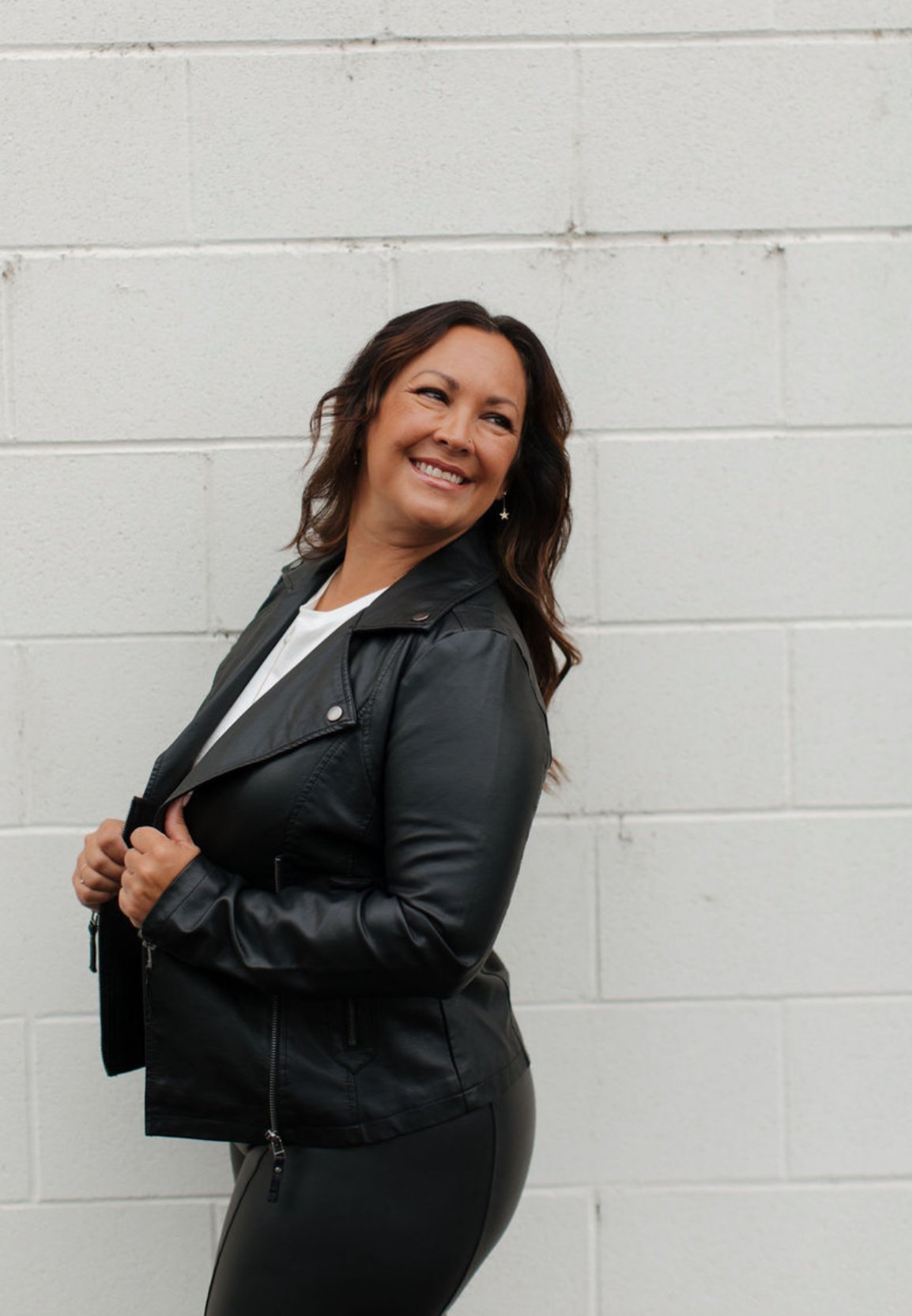 A woman with long wavy brown hair smiling while wearing a black leather jacket and black pants, standing against a white brick wall.