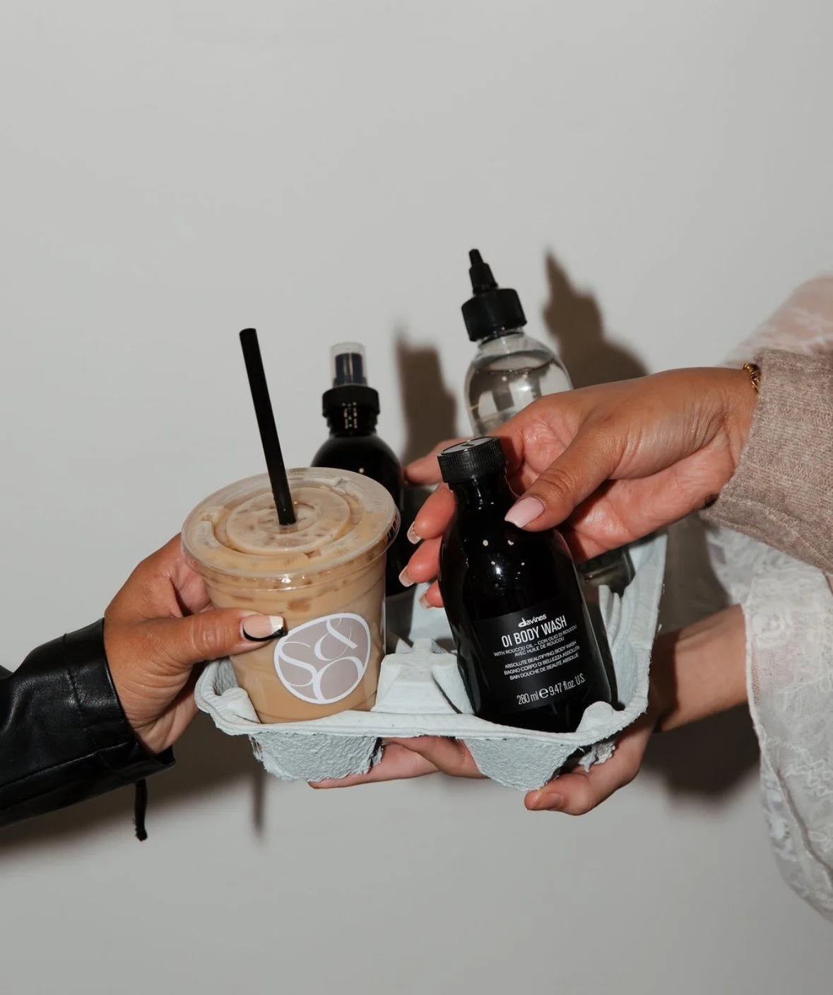 A person handing over a tray with skincare products and an iced coffee to another person. The tray holds a black bottle labeled 'Oi Body Wash' and other skincare bottles.