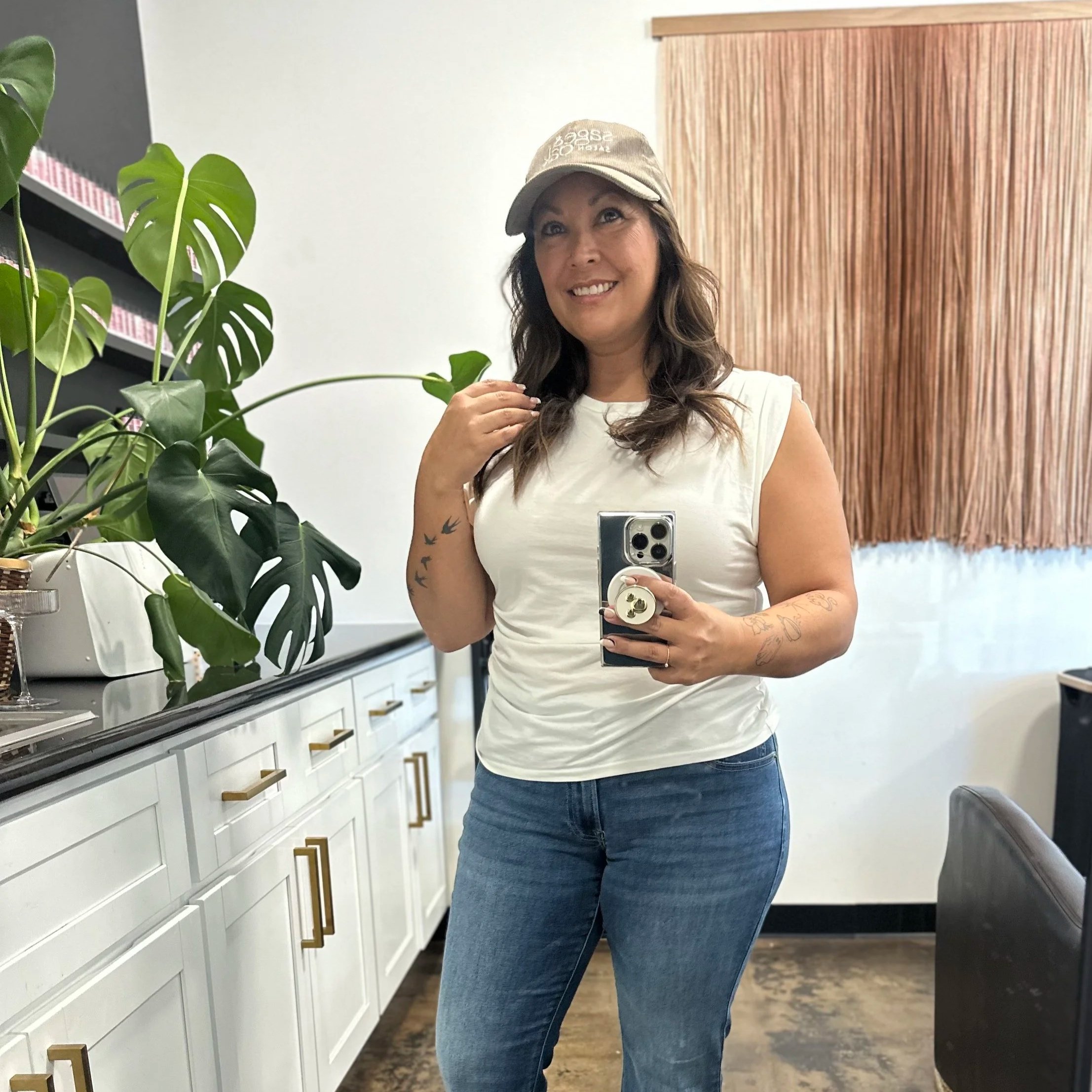 A woman taking a selfie in a room with white cabinets and a large green houseplant, wearing a beige cap and a white sleeveless top, with tattoos on her left arm.