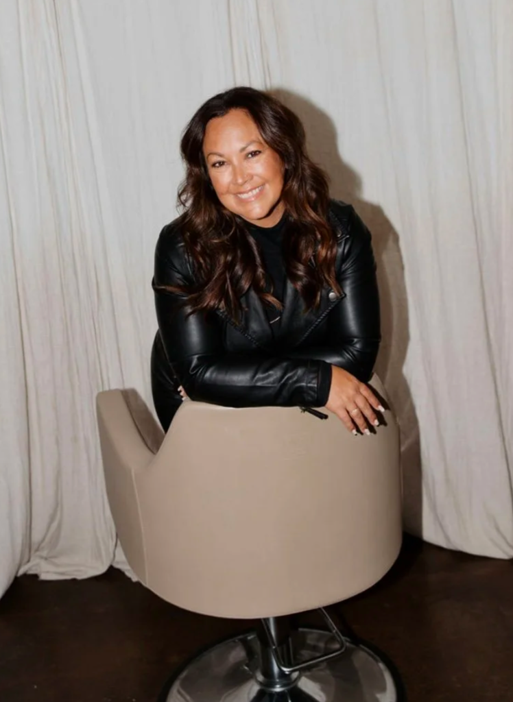 A woman with long brown hair, smiling, wearing a black leather jacket, leaning on the back of a beige salon chair.