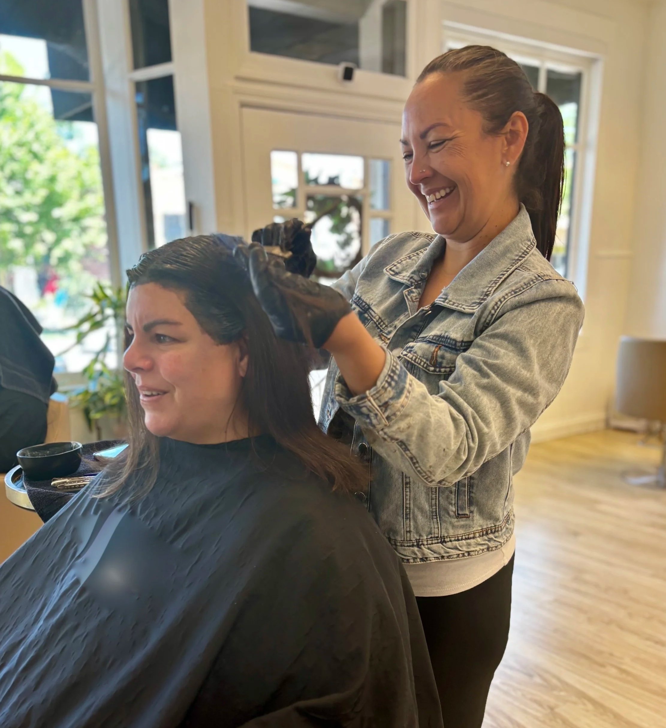 Woman with long hair smiling as a hairstylist applies hair dye or treatment, inside a bright, modern salon with large windows and wooden flooring.
