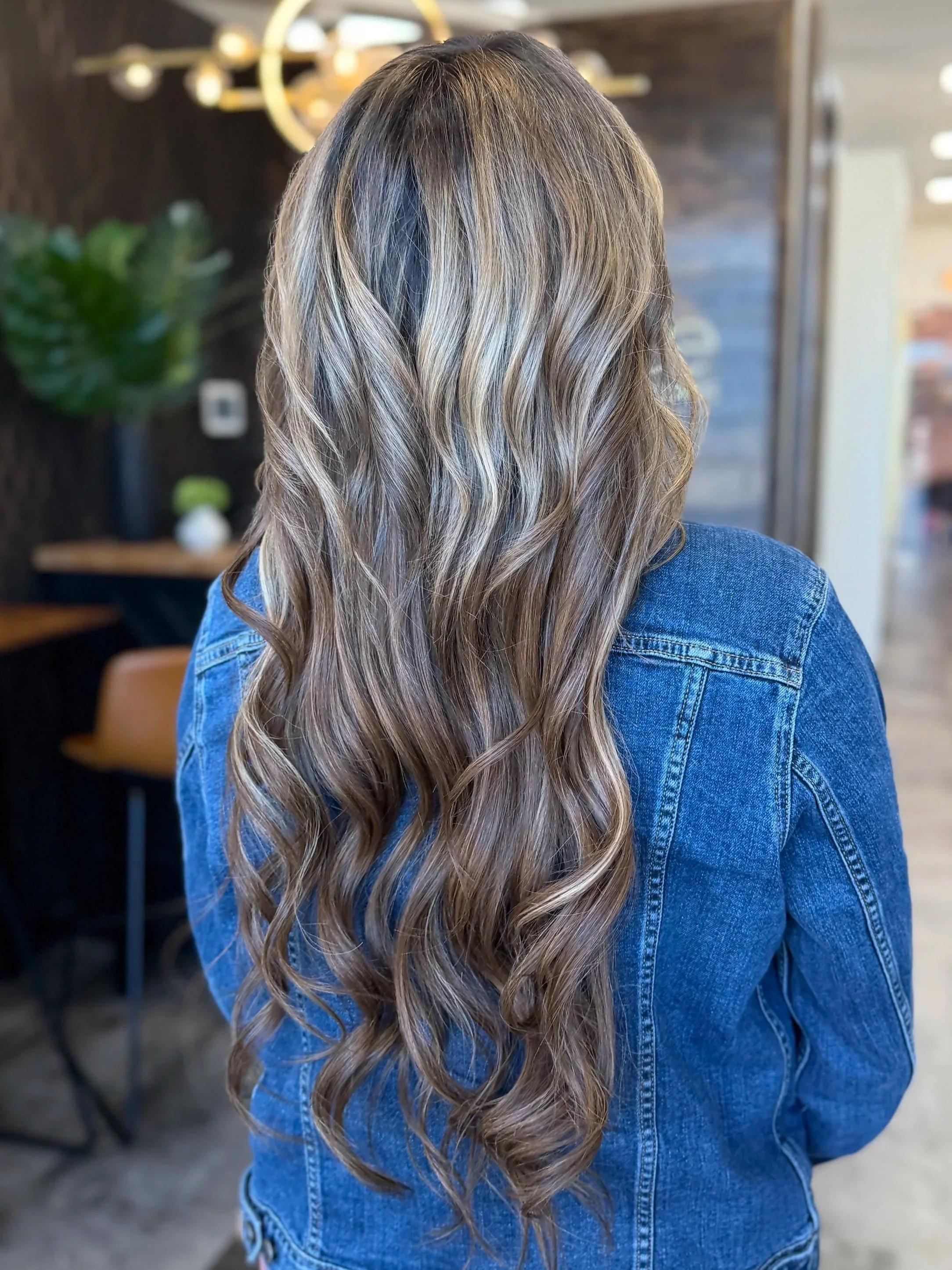 Back view of a woman with long, wavy, blonde hair wearing a denim jacket in a modern indoor space with dark walls, a plant, and decorative lighting.