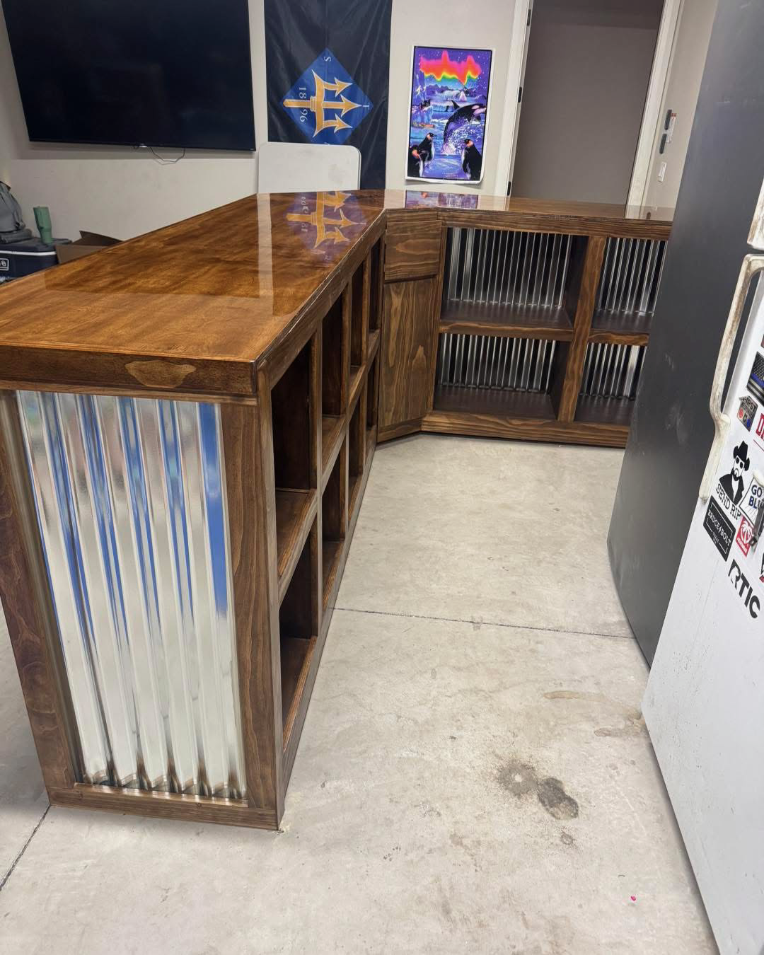 Wooden bar counter with an open shelf and corrugated metal accents, situated in a room with a television, artwork, and refrigerator.
