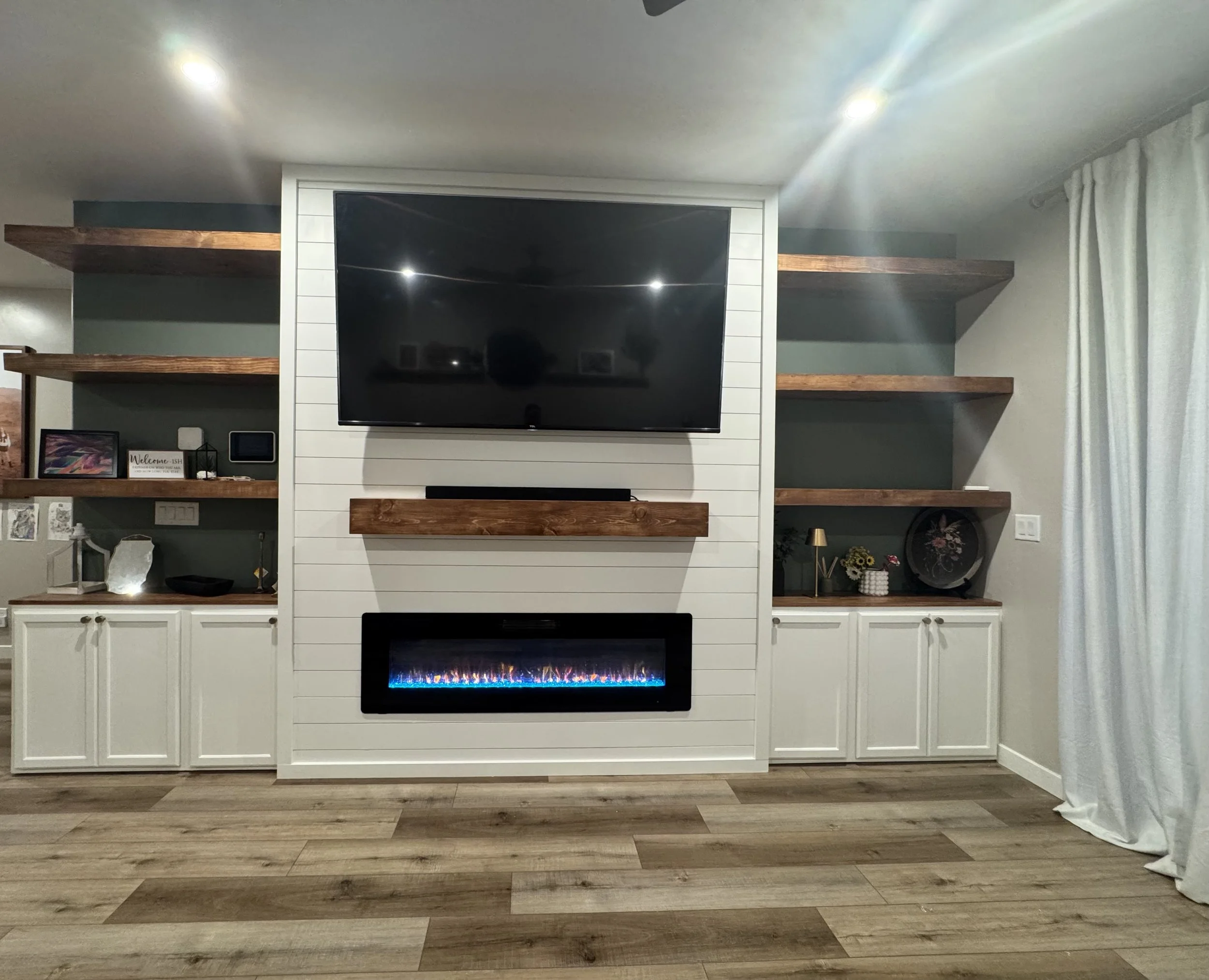 Living room with a large flat-screen TV mounted on a white shiplap wall, shelves with decorative items, and an electric fireplace below the TV. Hardwood floors and curtains on the right side.