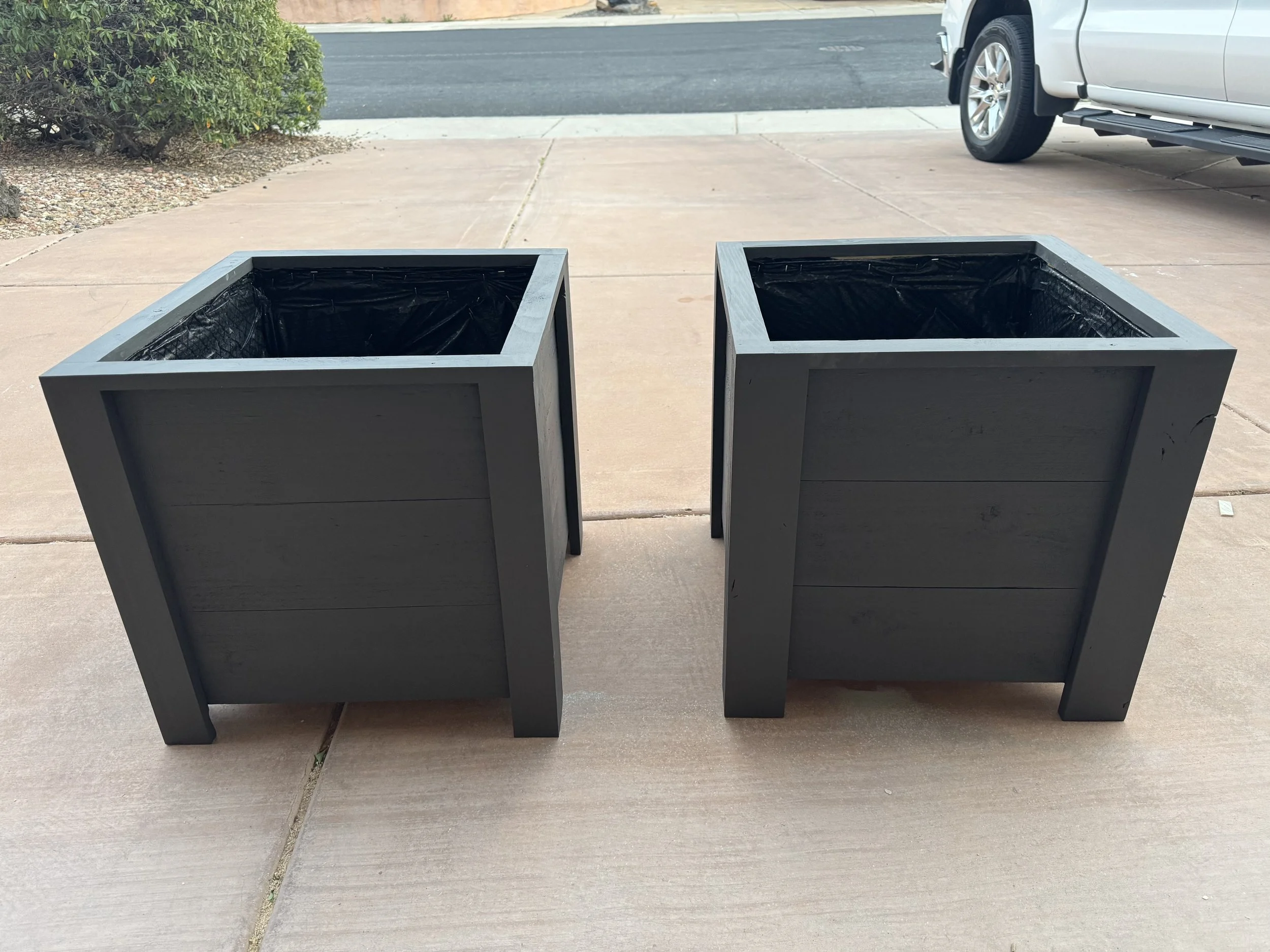 Two empty square planters with black linings on a concrete sidewalk in front of parked vehicles and a bush.