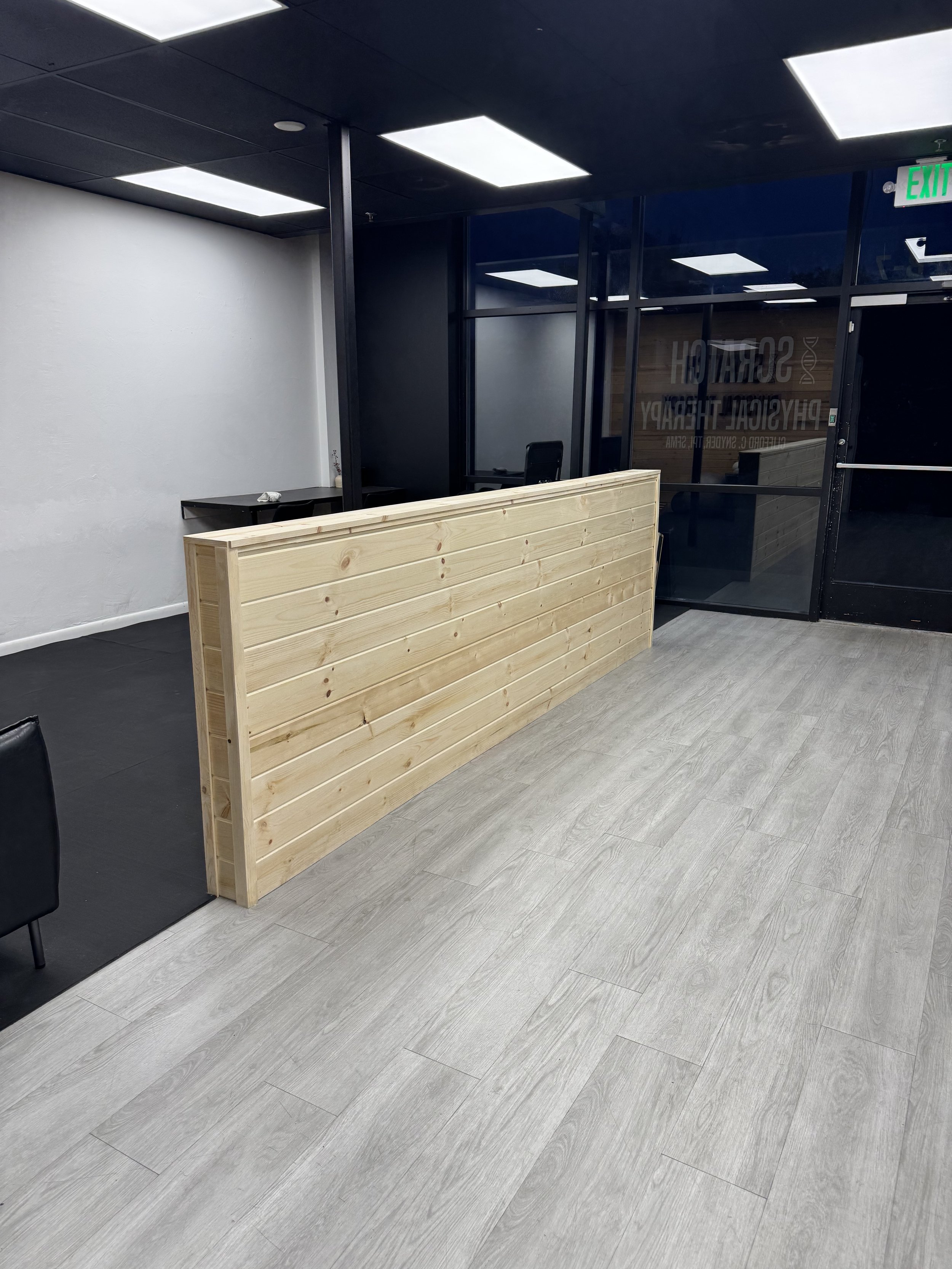 Interior of a modern office or clinic with grey wood flooring, a light grey wall, dark ceiling with rectangular fluorescent lights, a wooden reception counter, and black chairs. Glass walls with writing about a surgical and psychological therapy office in the background, an exit sign is visible.
