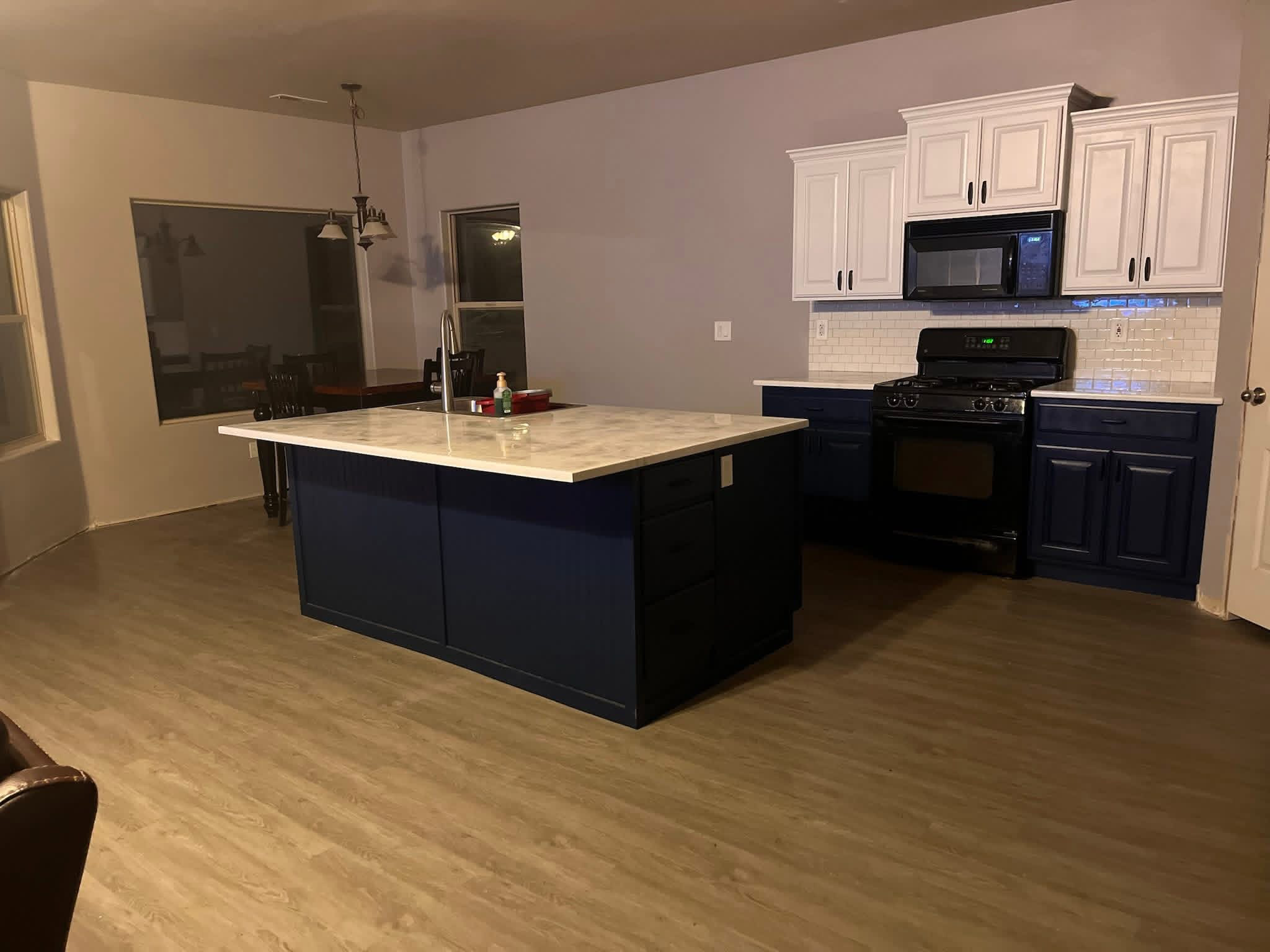 Empty kitchen with white upper cabinets, dark blue lower cabinets, black stove, microwave, beige tile backsplash, marble kitchen island, and wood flooring.