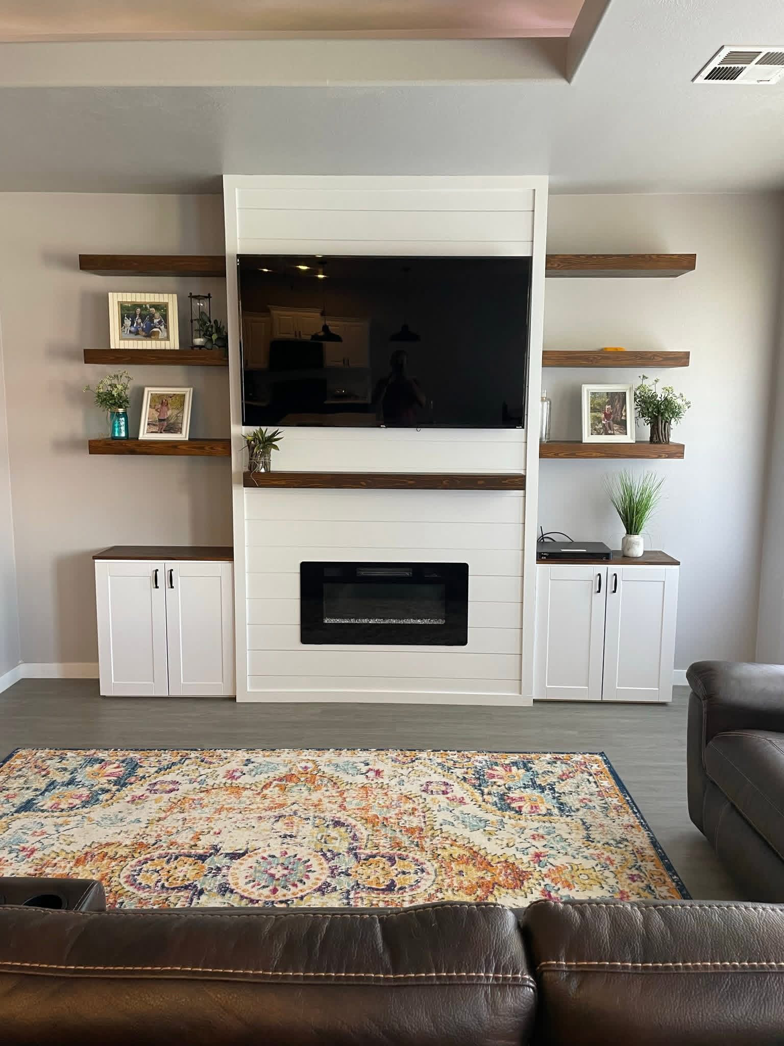 Living room with a white entertainment center, a black flat-screen TV, wooden shelves with framed photos and plants, a patterned area rug, and a brown leather couch.