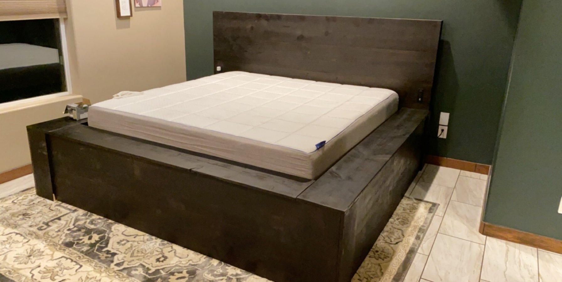 A wooden bed frame with a mattress on top, situated in a room with green and beige walls, a patterned rug, and tiled flooring.