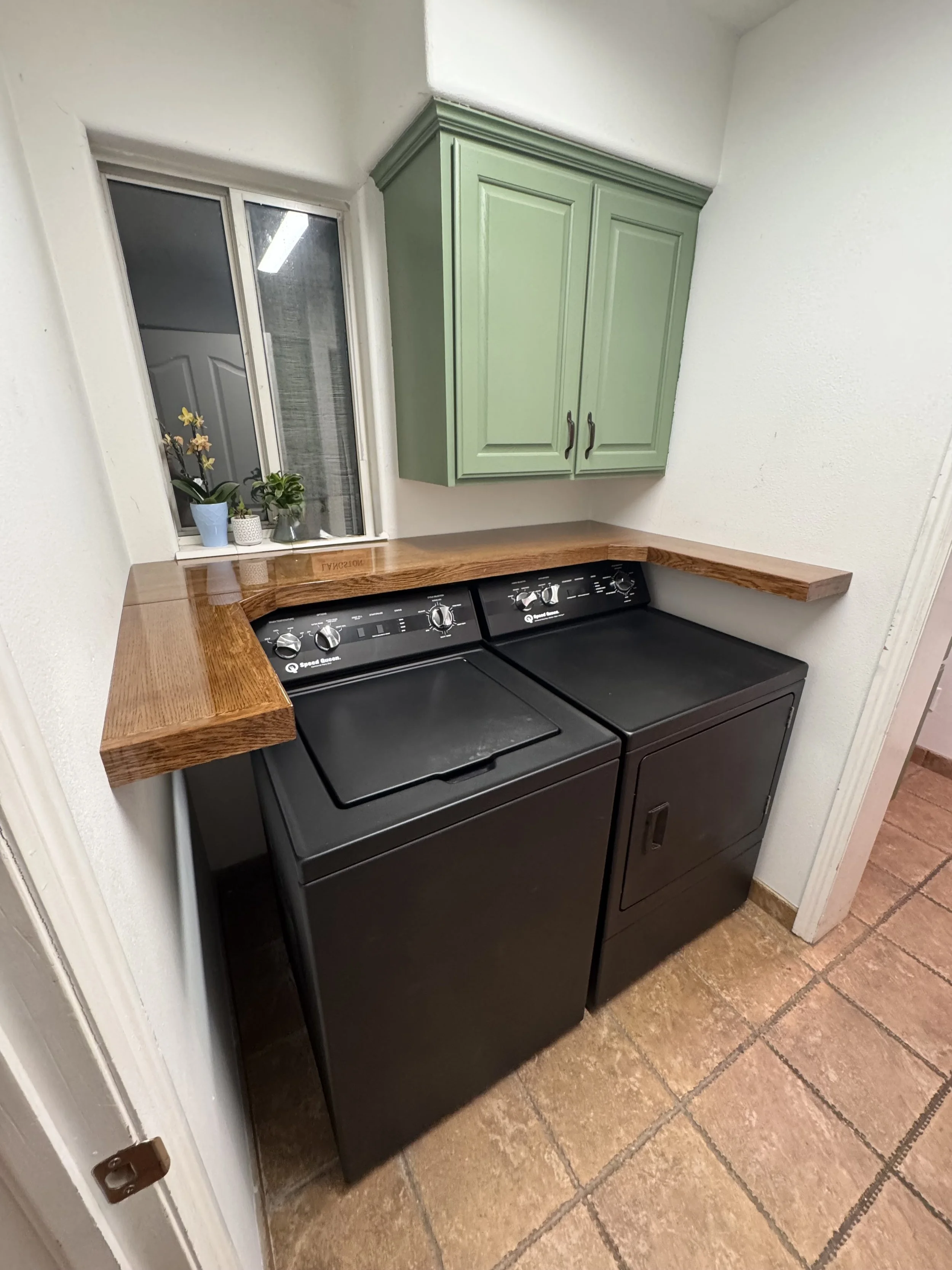 A laundry area with a black washing machine, a wooden countertop, a green wall-mounted cabinet, a window with potted plants on the windowsill, and a tiled floor.