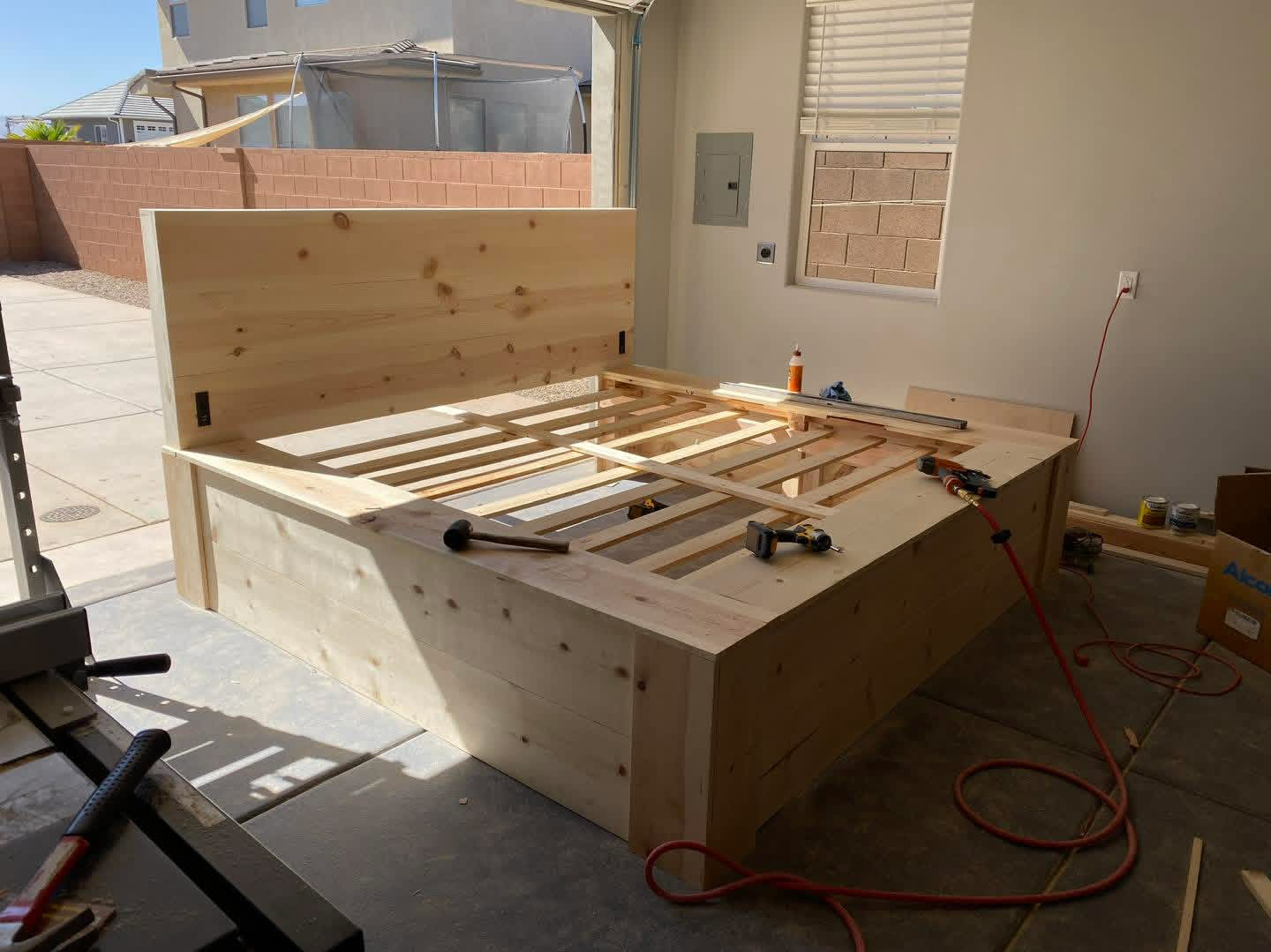 Wooden bed frame under construction inside a garage or workshop. Tools, glue, and supplies are on the bed frame and nearby surfaces. A window and wall with electrical panel are visible.