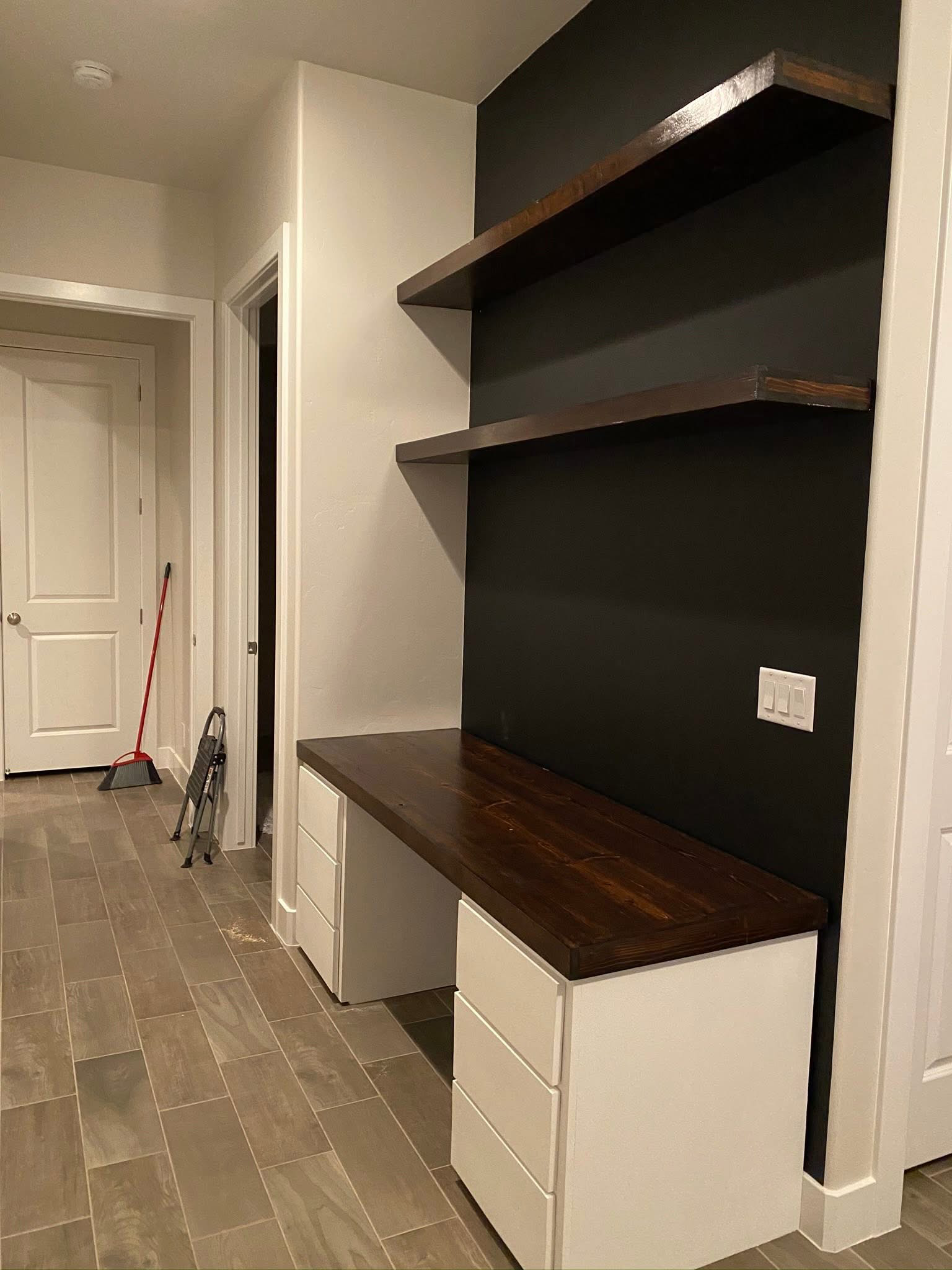 Interior view of a modern kitchen or dining area corner with white cabinetry, a wooden countertop, and dark floating shelves. There's a white wall with a double light switch, and the floor is tiled with brownish tiles. A mop and a step ladder are leaning against the wall near the door.