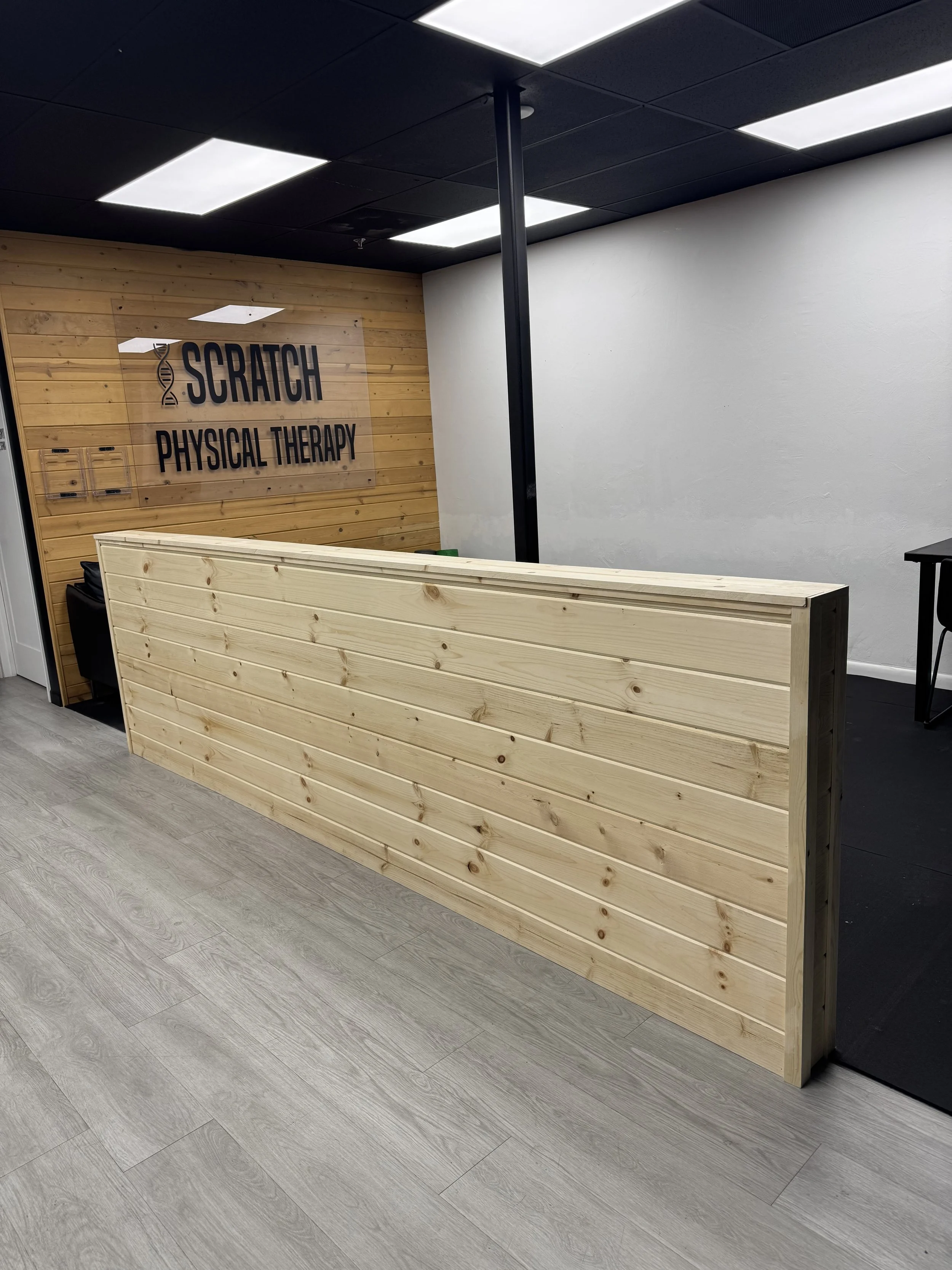 Reception area at SCRATCH PHYSICAL THERAPY with a wooden desk, black chairs, and a wooden wall with the business name and logo.