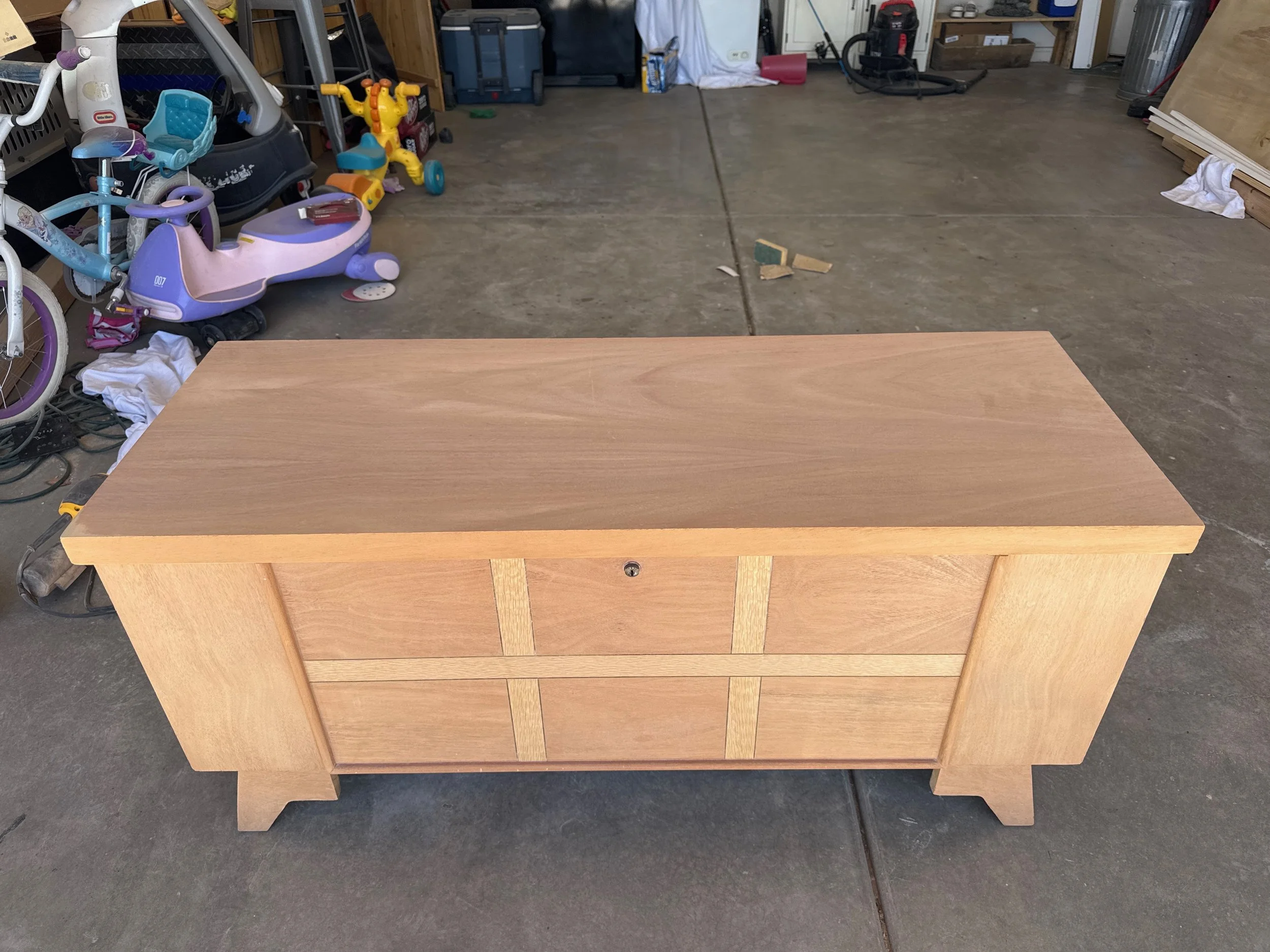 A light-colored wooden dresser with four drawers in a garage or workshop setting. The top drawer has a keyhole, and the dresser has a simple, sturdy design. In the background, various toys, tools, and storage items are visible.