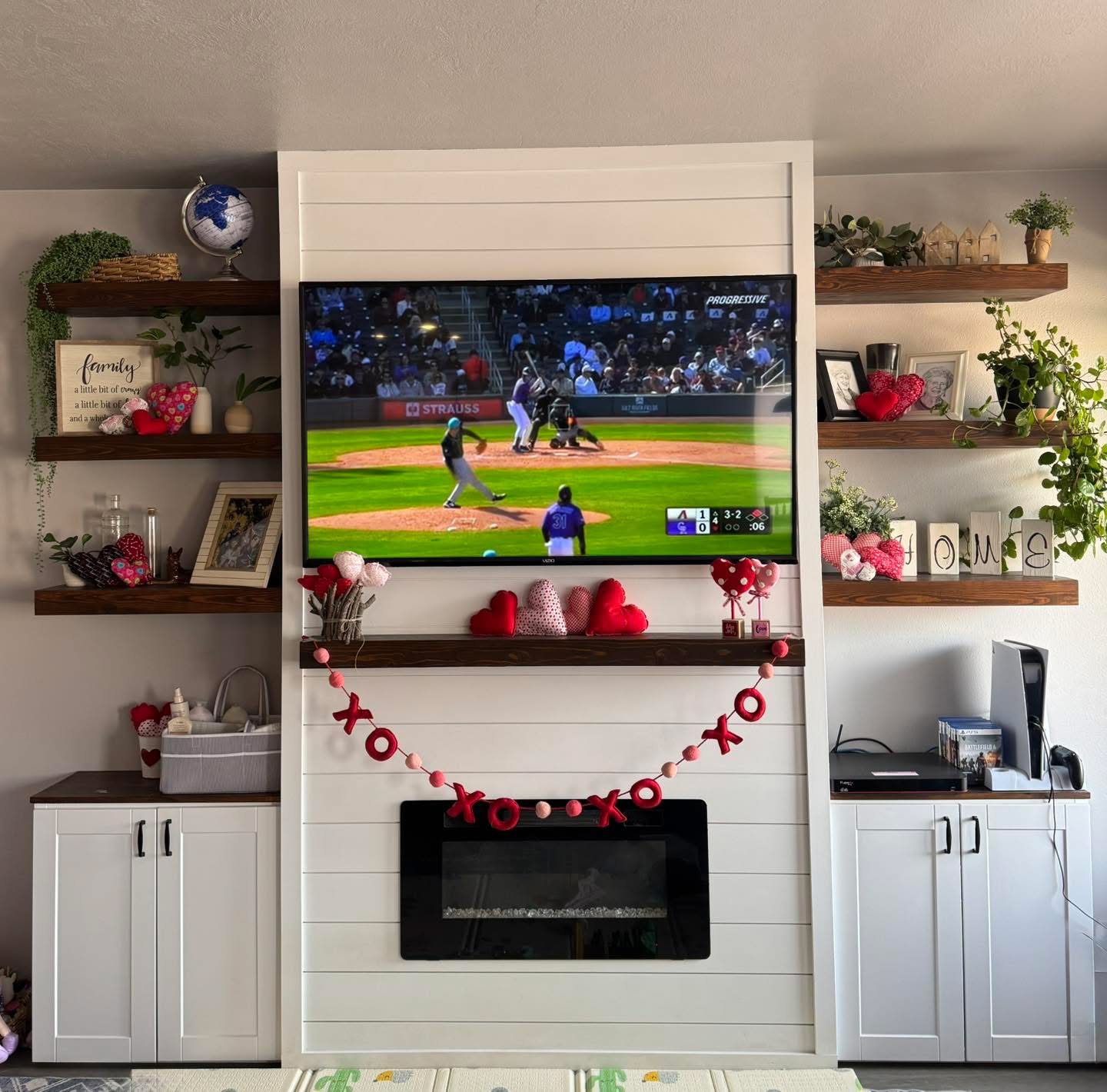 Living room with a white fireplace, decorated for Valentine's Day, with a large TV showing a baseball game, surrounded by shelves with picture frames, plants, and decorative items, including hearts and the word "love".