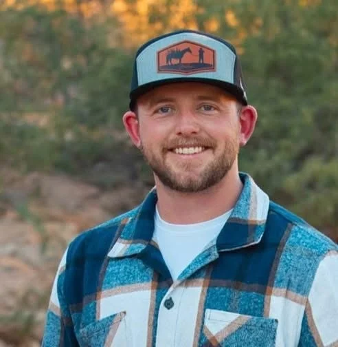 A man outdoors wearing a blue plaid shirt and a trucker hat with a mountain and animal logo, smiling with a background of trees.