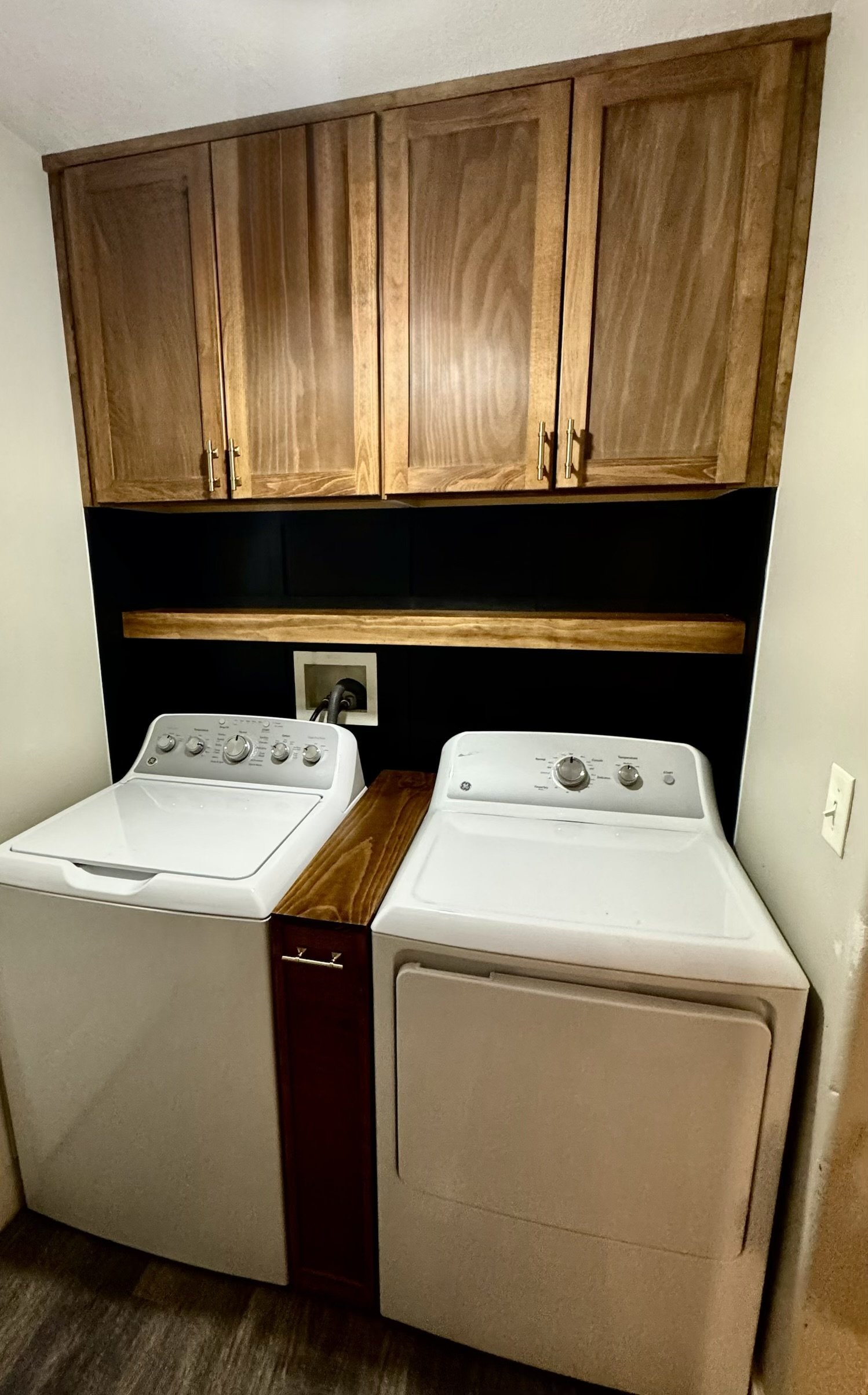 A laundry room with a washer and dryer side by side, mounted on a dark wood cabinet, with wooden wall cabinets above for storage.