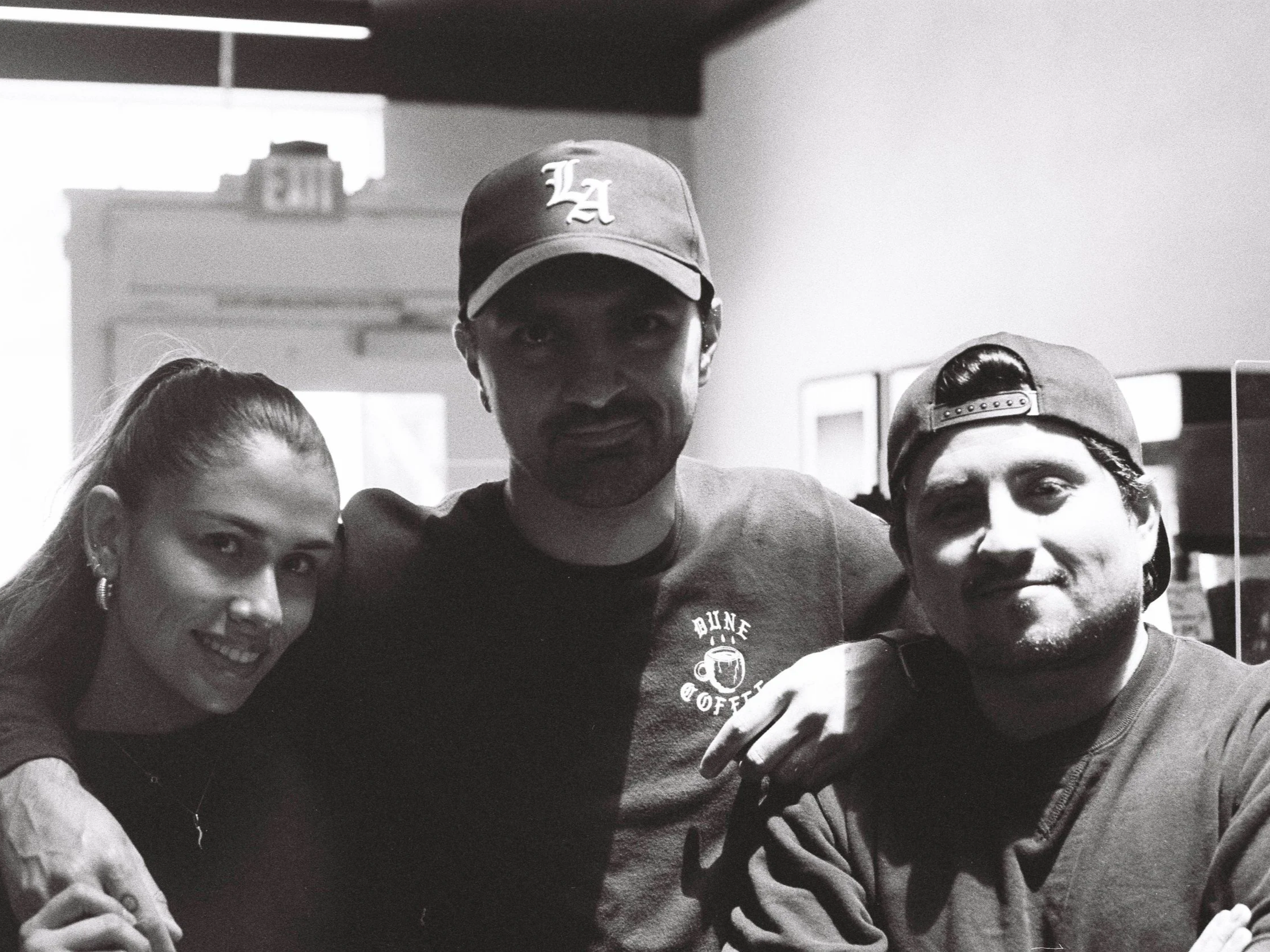 A black-and-white photo of three young people, two women and one man, posing closely together in an indoor setting. The woman on the left has her hair tied back and is smiling. The man in the middle wears a cap with