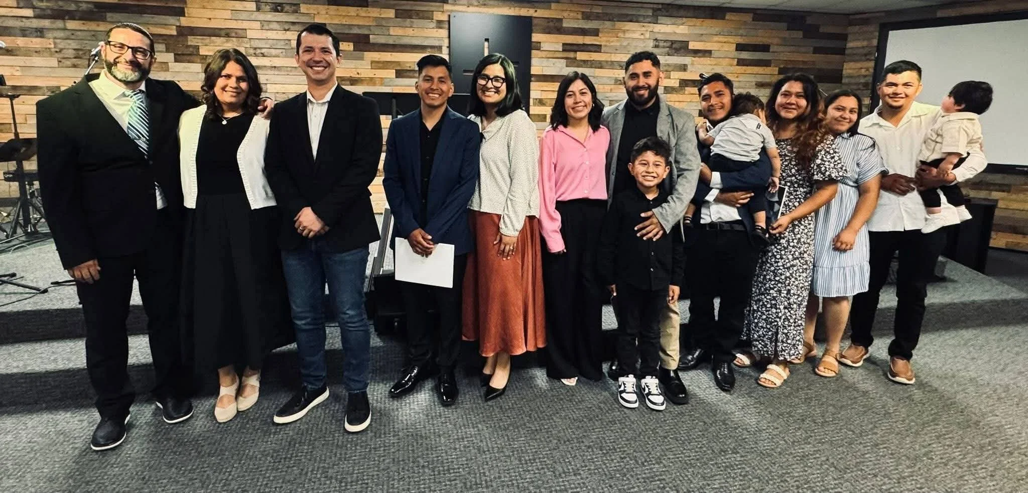 Group of people standing together indoors, smiling, in front of a wooden wall, some holding children.