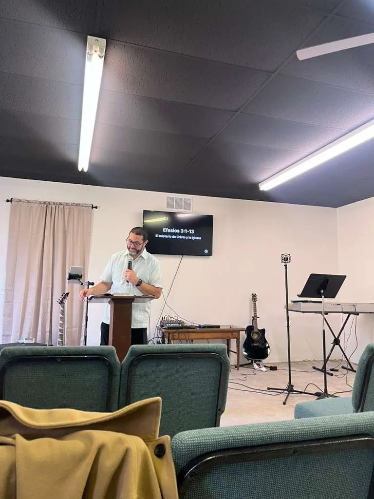 Man giving a lecture or sermon at a church or community center, with musical instruments like a guitar, keyboard, and microphone behind him, and a television screen displaying a Bible verse in Spanish.