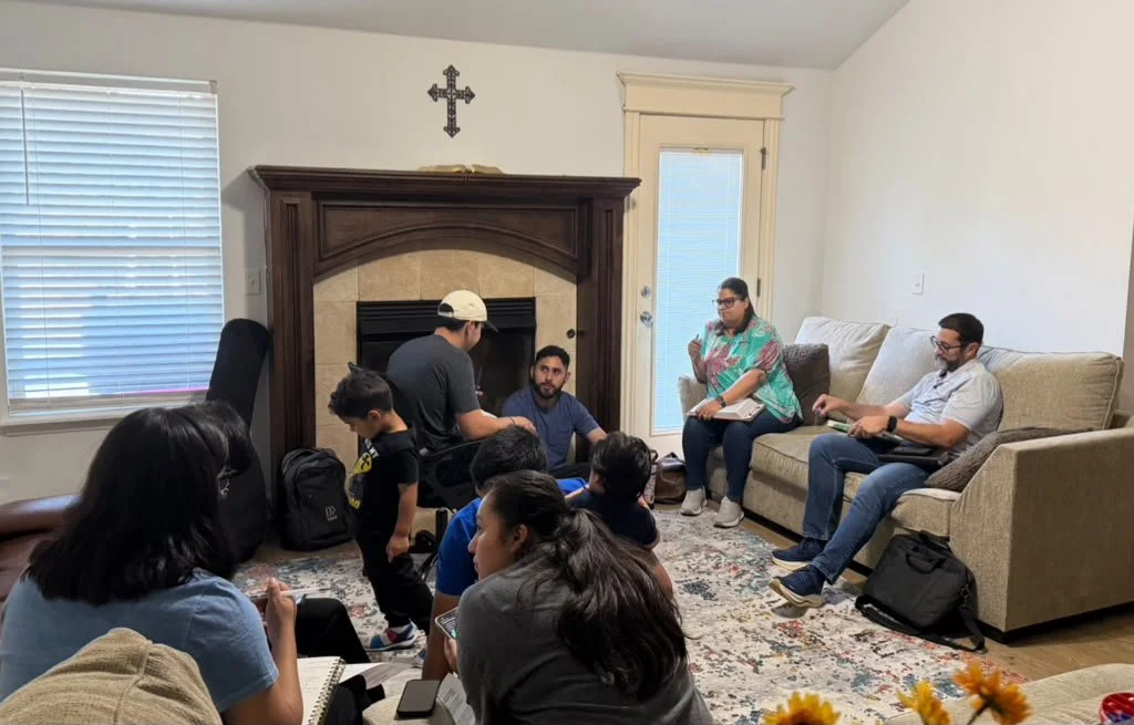 A group of people sitting and standing in a living room, engaged in a discussion or meeting. There are seven people visible, with two sitting on a couch, three sitting on the floor, and two standing near the fireplace. The room has a window with blinds, a fireplace, and a door leading outside. A cross hangs above the fireplace.