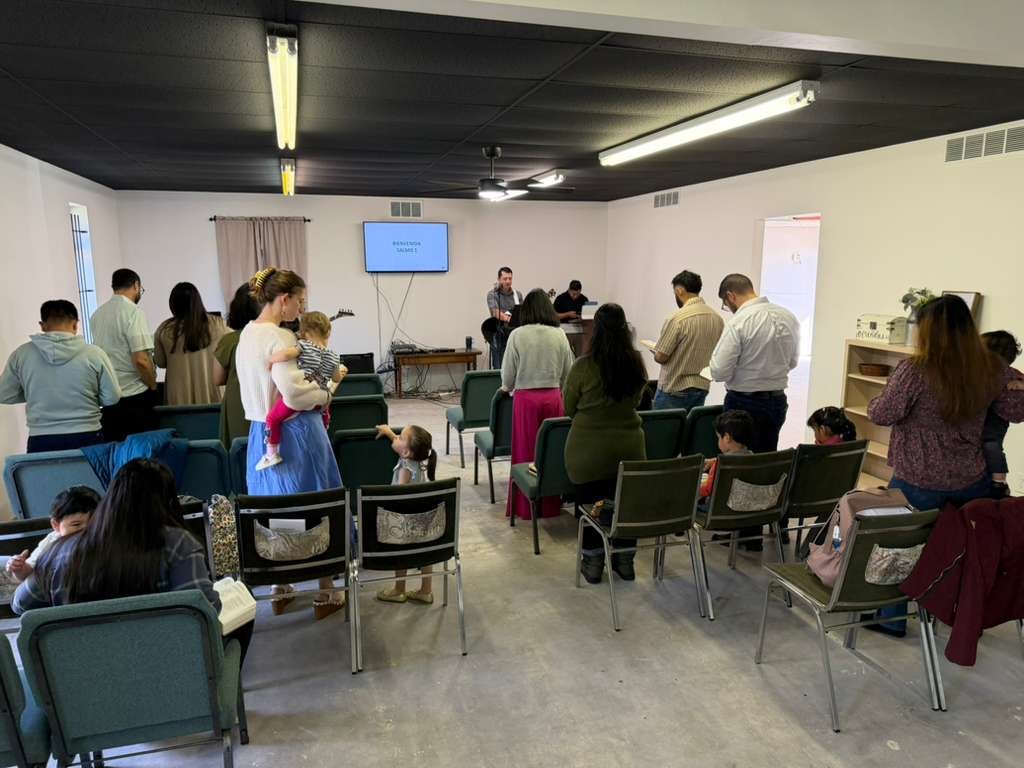 People gathered in a small worship service or prayer meeting, with some standing and others sitting in chairs, in a room with plain white walls, a flat-screen TV, and a music stand at the front.