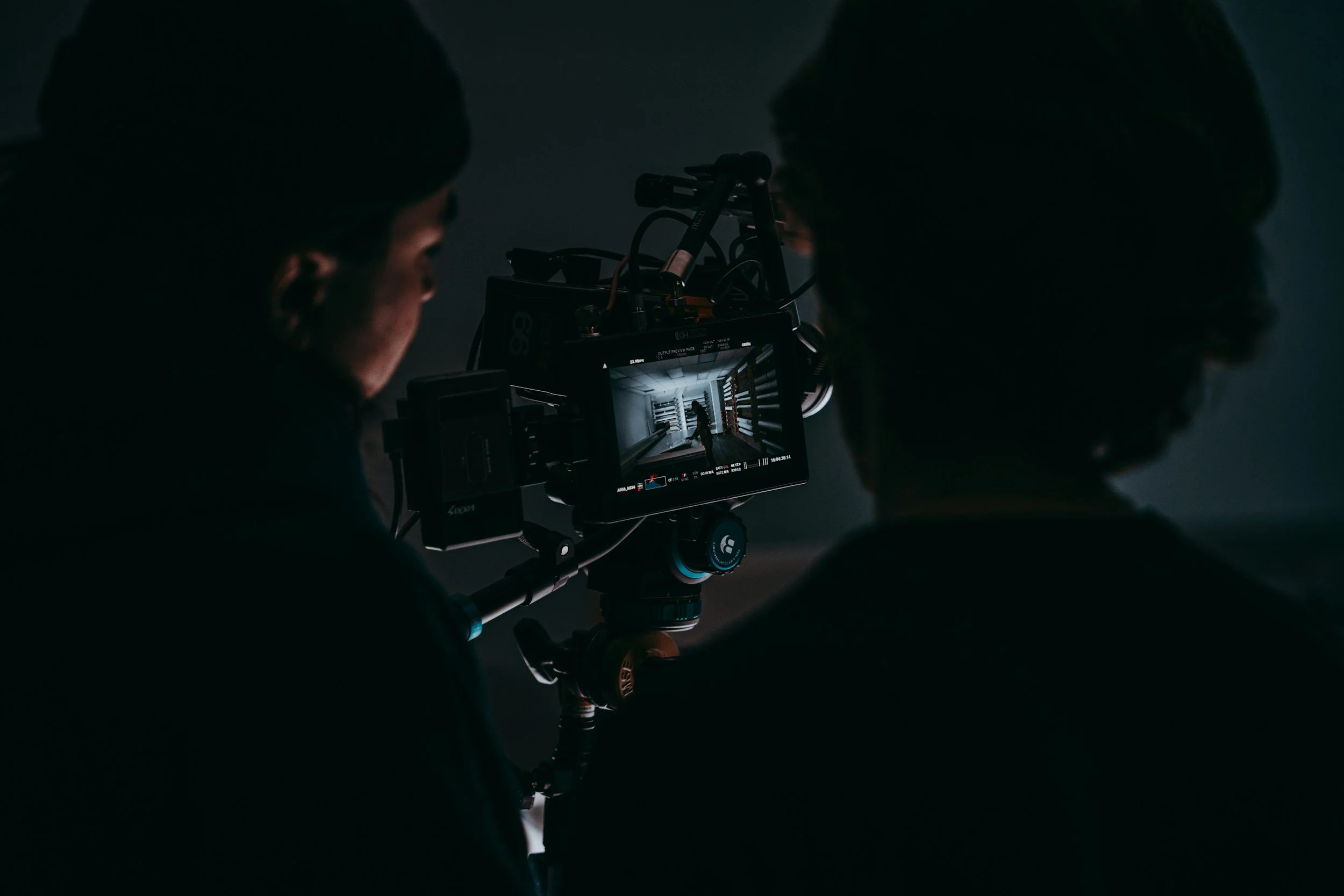 Two individuals operate a professional camera on a tripod in a dark room, capturing footage of a corridor with shelves.