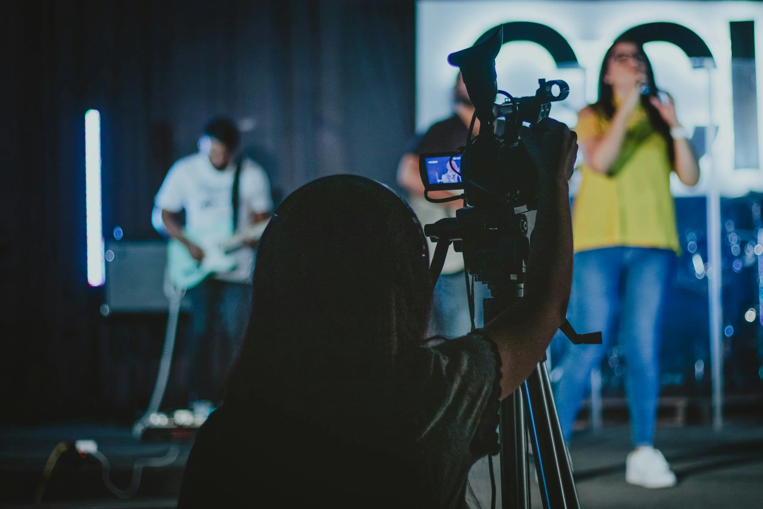 A person operating a professional camera captures a scene of a woman talking into a microphone and two men, one playing a guitar, on a stage in a dimly lit studio or event space.
