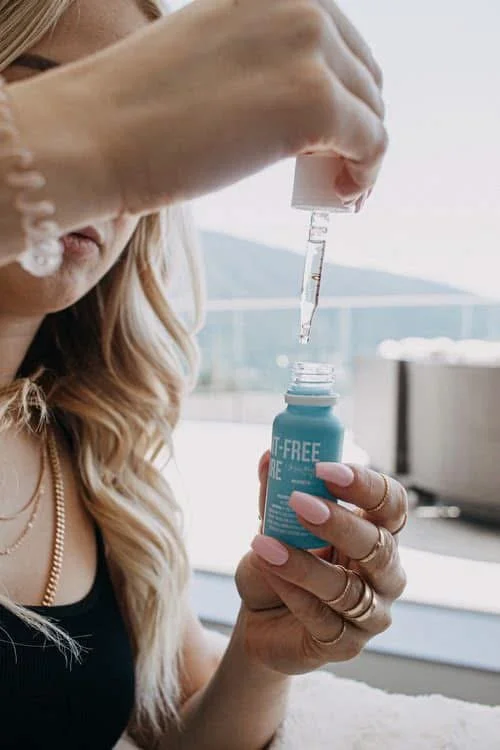 A woman using a dropper to transfer liquid into a small container with a label, likely in a setting with natural light and a view of the outdoors.
