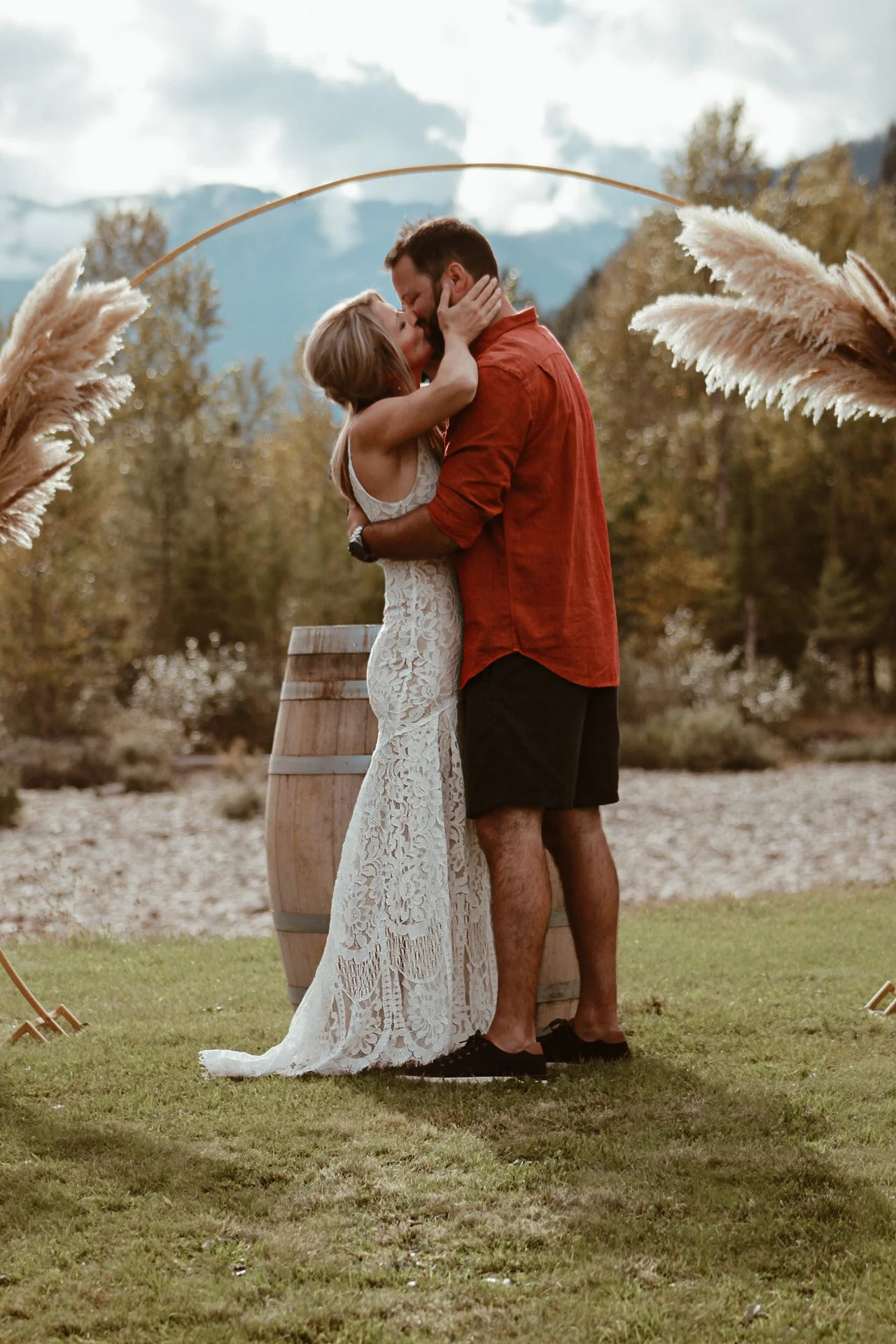 A couple is kissing outdoors in front of a wine barrel with a scenic mountain view and cloudy sky in the background. The woman is wearing a white lacy dress, and the man is wearing a red shirt and black shorts. Pampas grass decorates the scene.