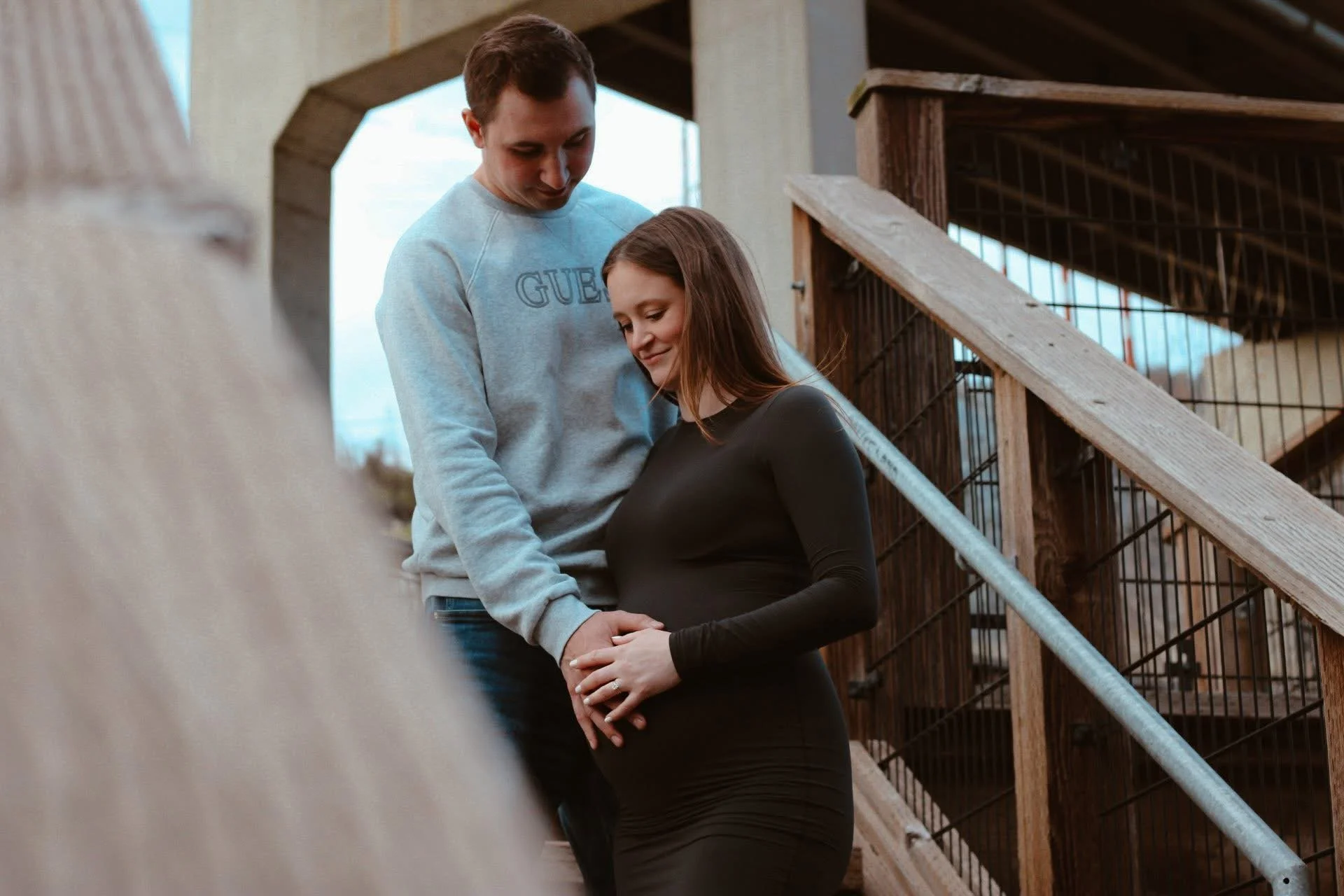 A young man and a pregnant woman standing close together on a staircase outdoors, holding hands and smiling gently, with a wooden staircase and structure in the background.