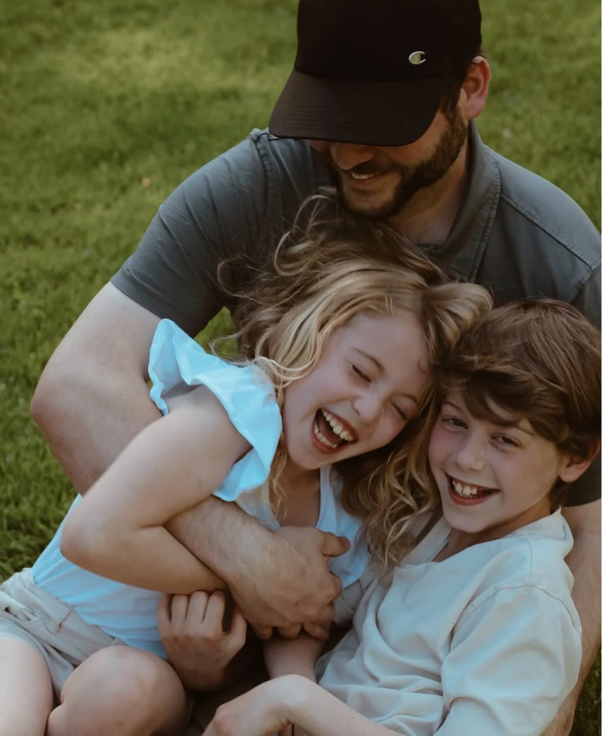 A man and two children are outdoors on green grass, hugging and laughing, appearing joyful and playful.