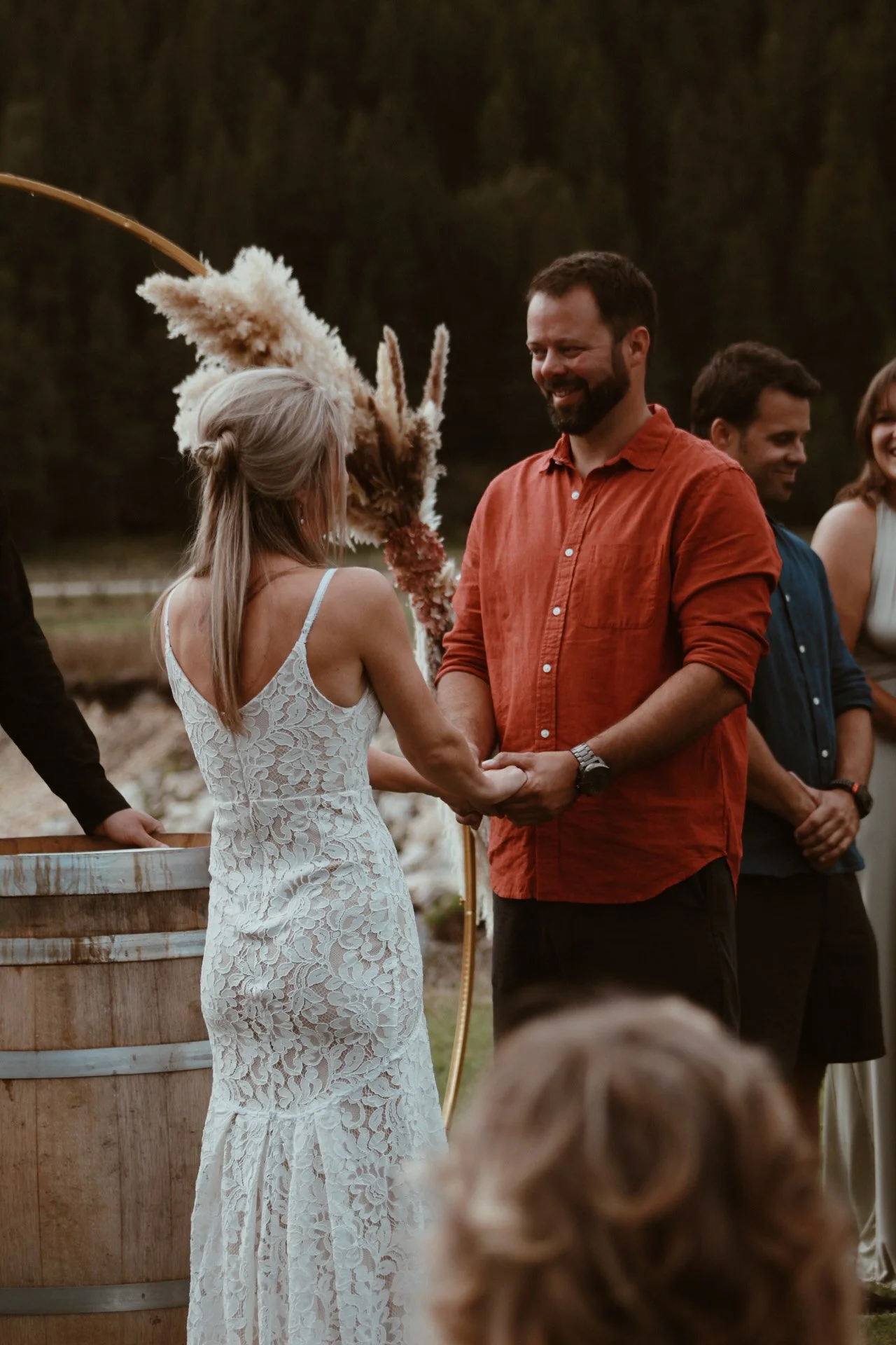 A wedding ceremony outdoors with a bride and groom holding hands, surrounded by friends and family. The bride wears a white lace dress, and the groom wears an orange shirt. There are tall grasses and trees in the background.