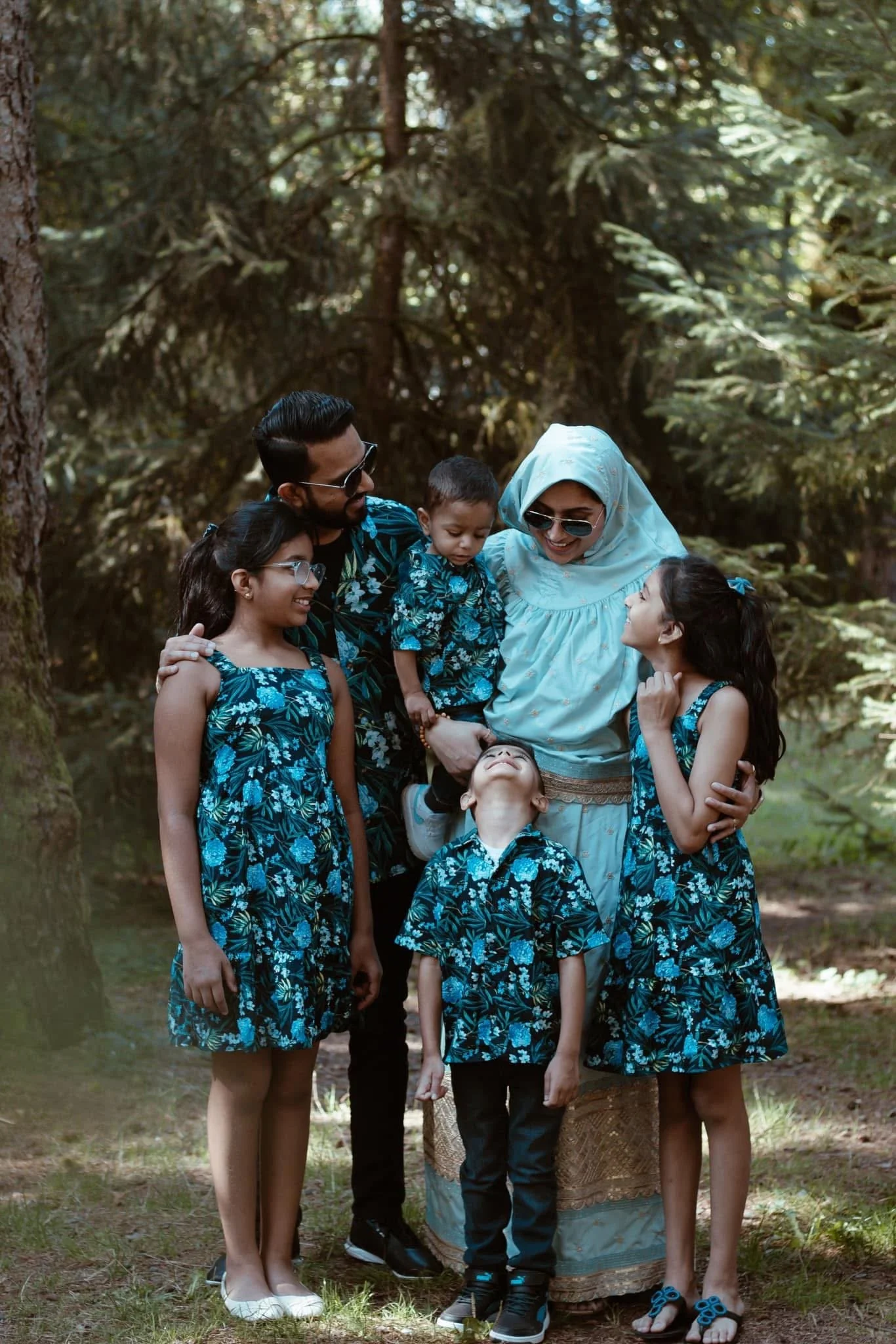 A family of six standing outdoors in a forest, smiling and looking at each other. The group includes a man, a woman wearing a headscarf, three girls, and two boys. All are dressed in matching blue floral clothing.
