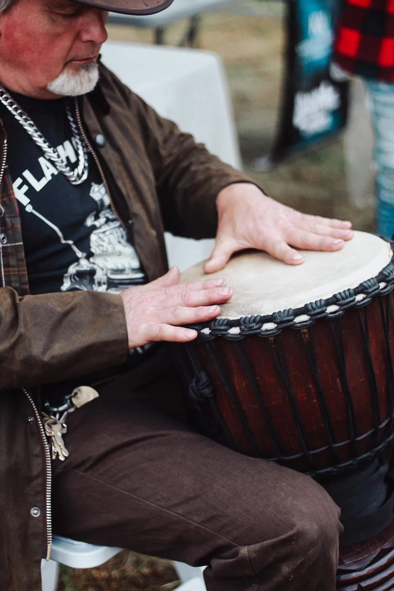 A person playing a percussion drum, possibly a conga, with hands on the drum surface. The person is wearing a brown leather jacket, dark pants, and a black T-shirt with a graphic print. The background includes another person in striped pants and a bo