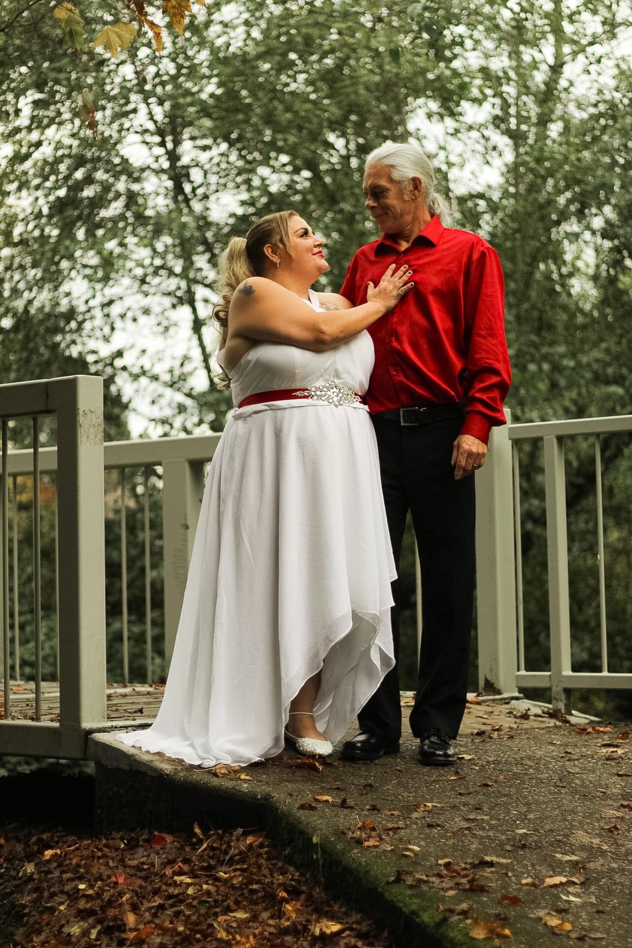 A woman in a white dress with a red waistband and a man in a red shirt share a moment on a bridge surrounded by fall foliage, looking into each other's eyes.