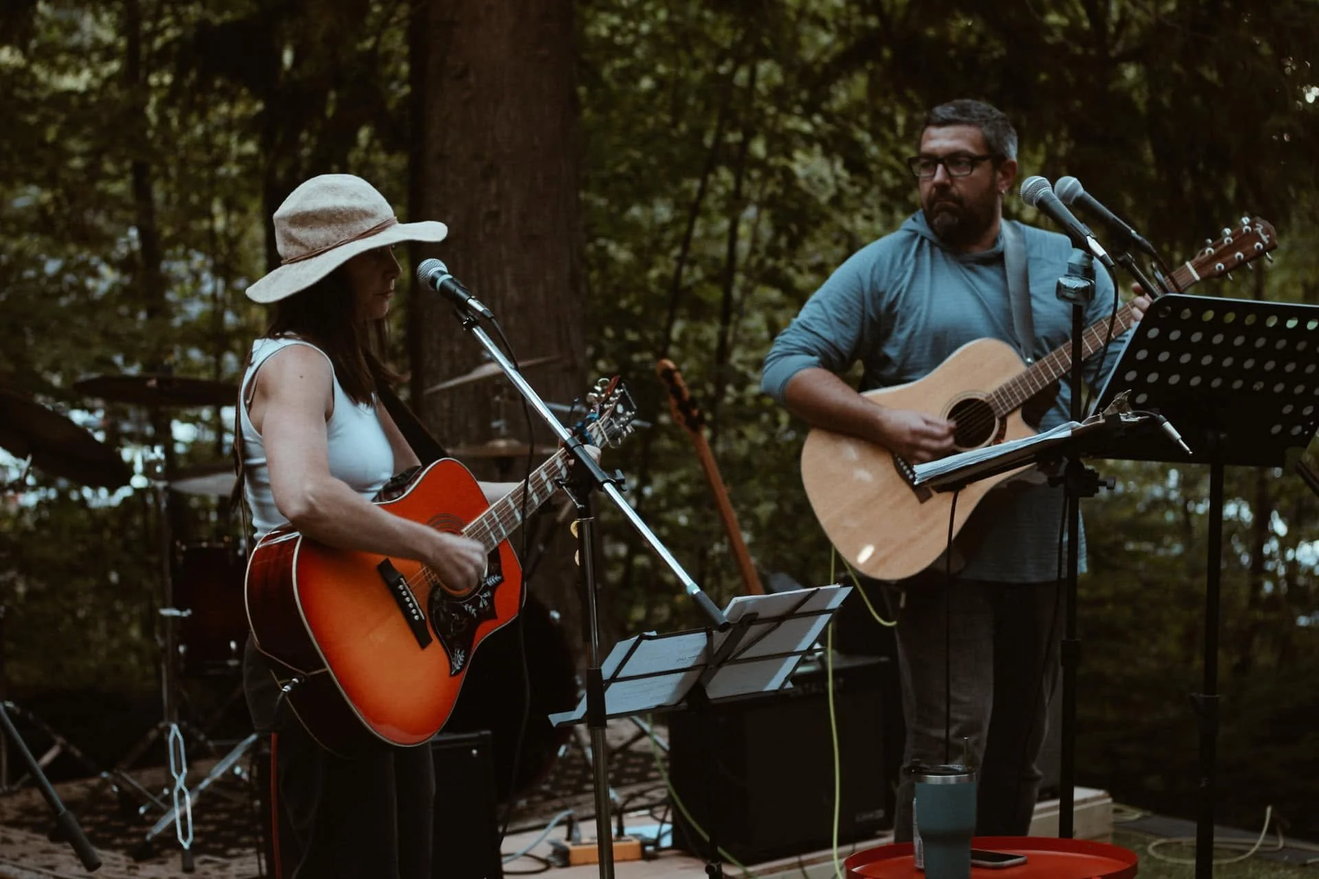 Two musicians playing guitars outdoors in a wooded park, one woman wearing a sun hat and the other man with glasses, with microphones and music stands around them.